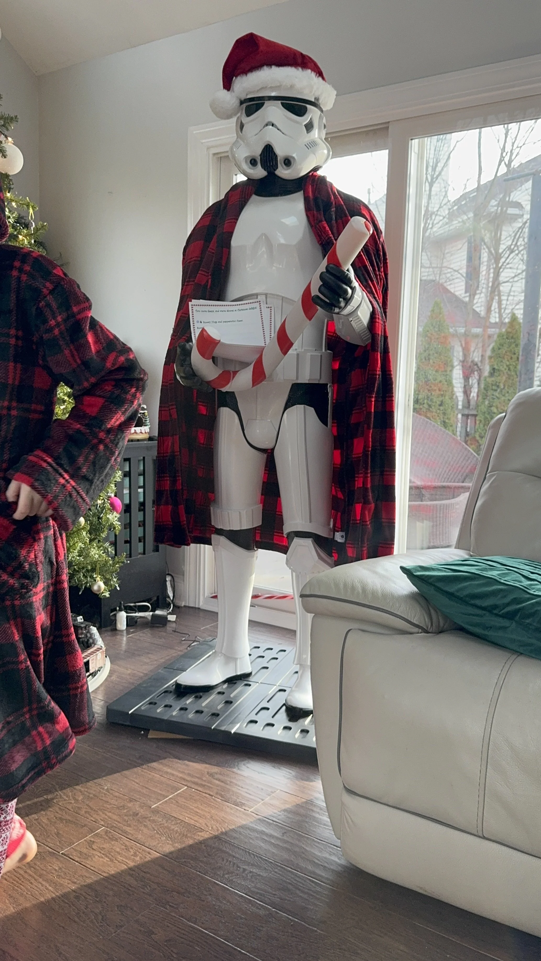 A life-sized robot dressed as Santa Claus, wearing a red Santa hat, a red plaid cape, and holding a candy cane, is standing inside a living room near a large window and a Christmas tree.