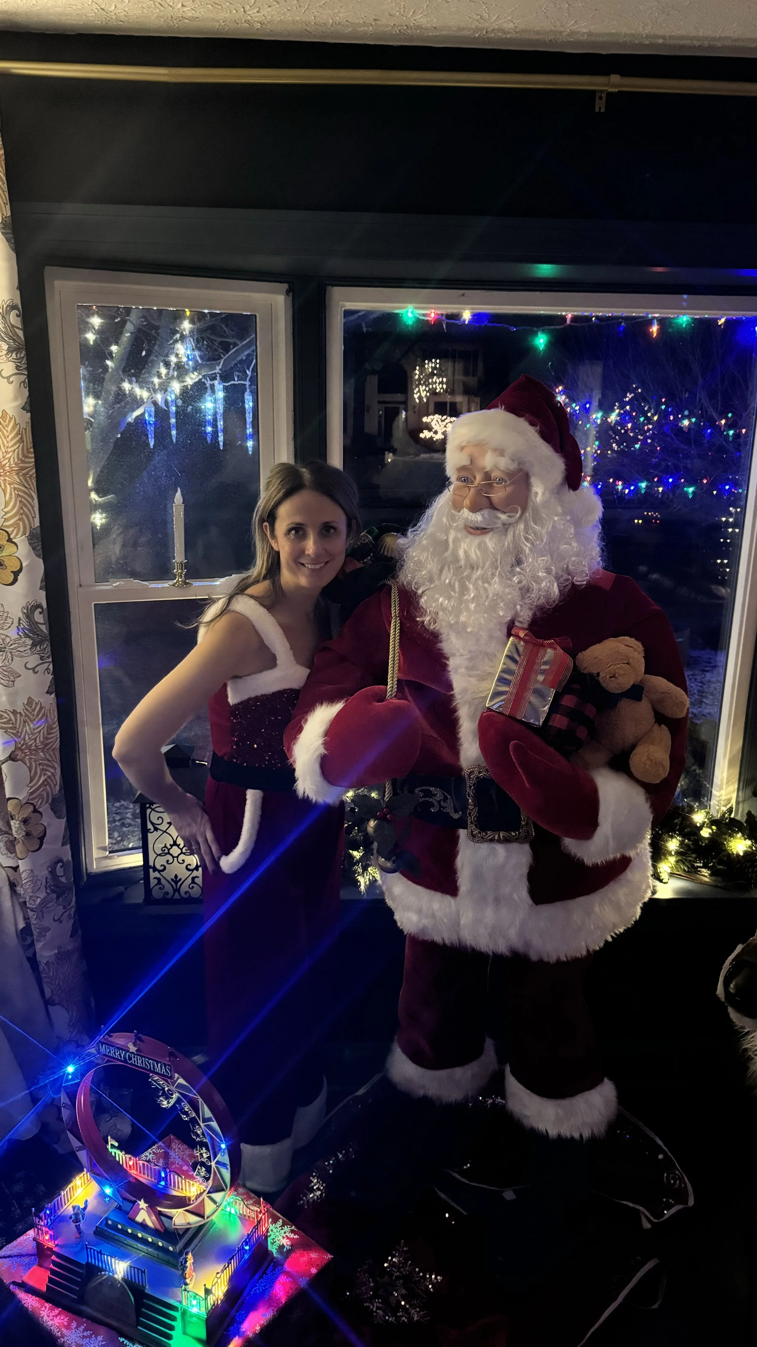 A woman in a Santa Claus dress stands next to an animatronic Santa Claus, posing for a photo in front of a window decorated with colorful Christmas lights. The woman has long brown hair and is smiling.