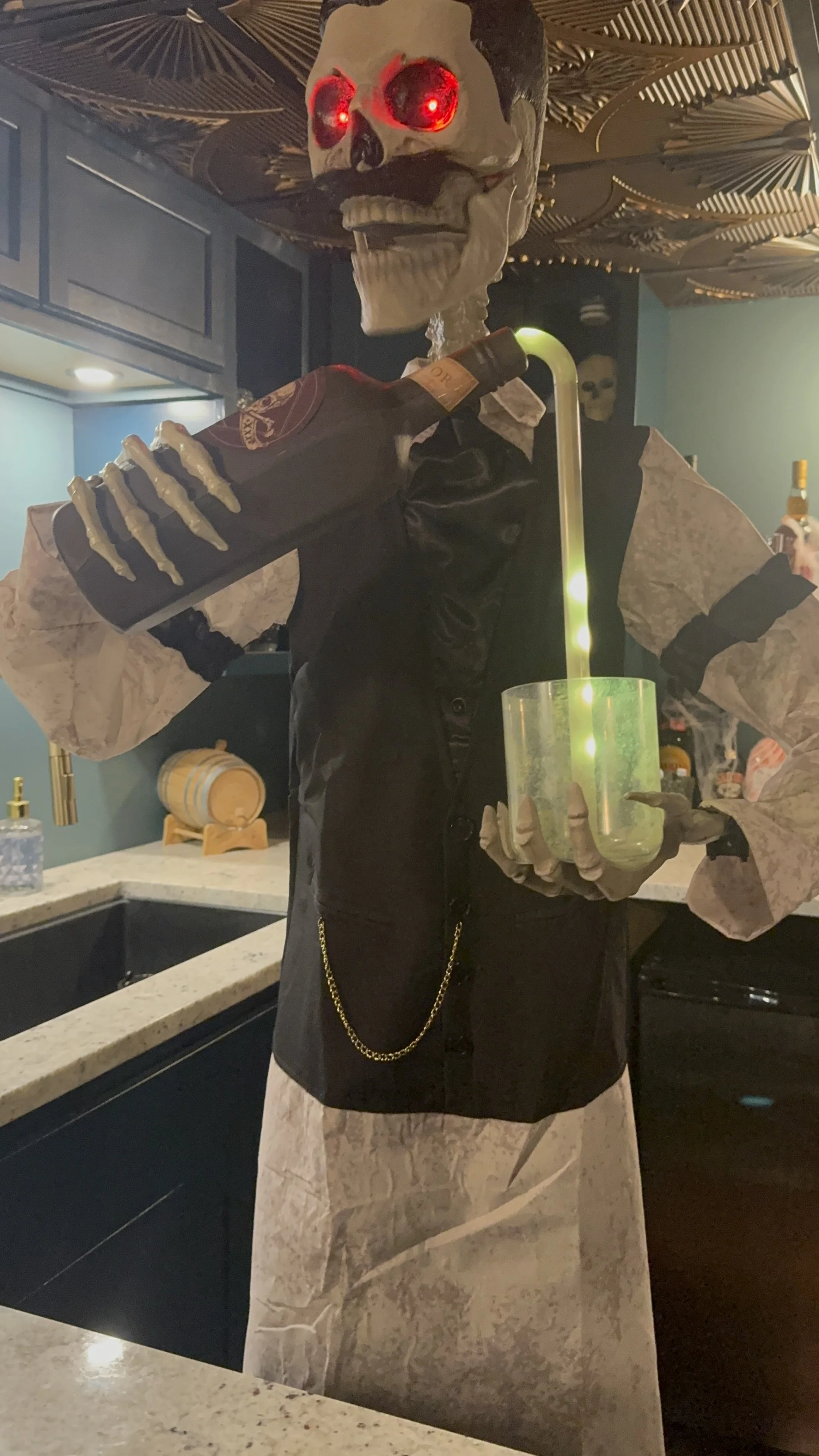 Animatronic  bar tender skeleton with glowing red eyes, holding a bottle and a glowing drink container, standing in a bar or restaurant setting.