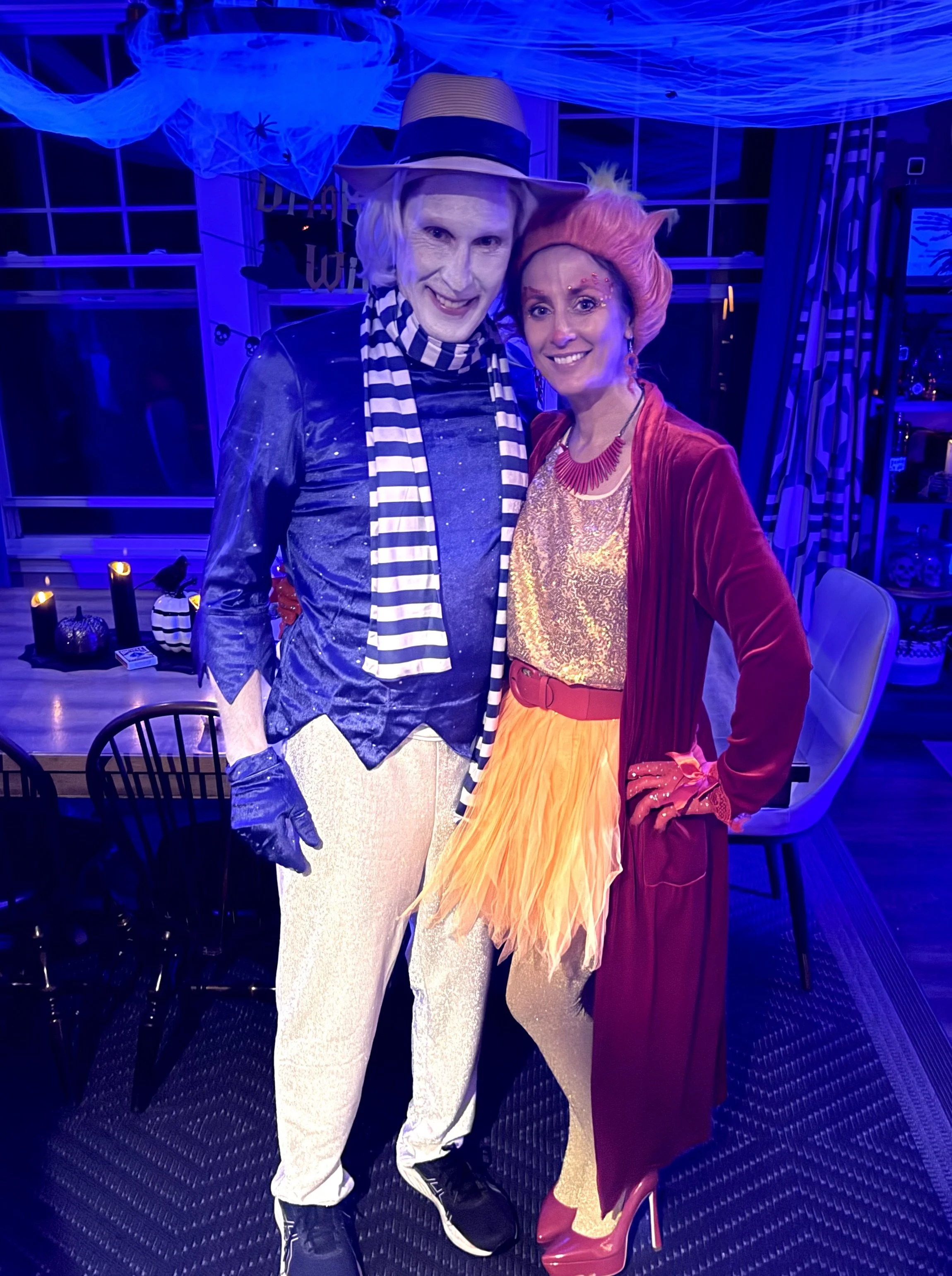 Two people dressed in colorful costumes standing close together at a Halloween or costume party, with decorations like cobwebs and candles in the background.