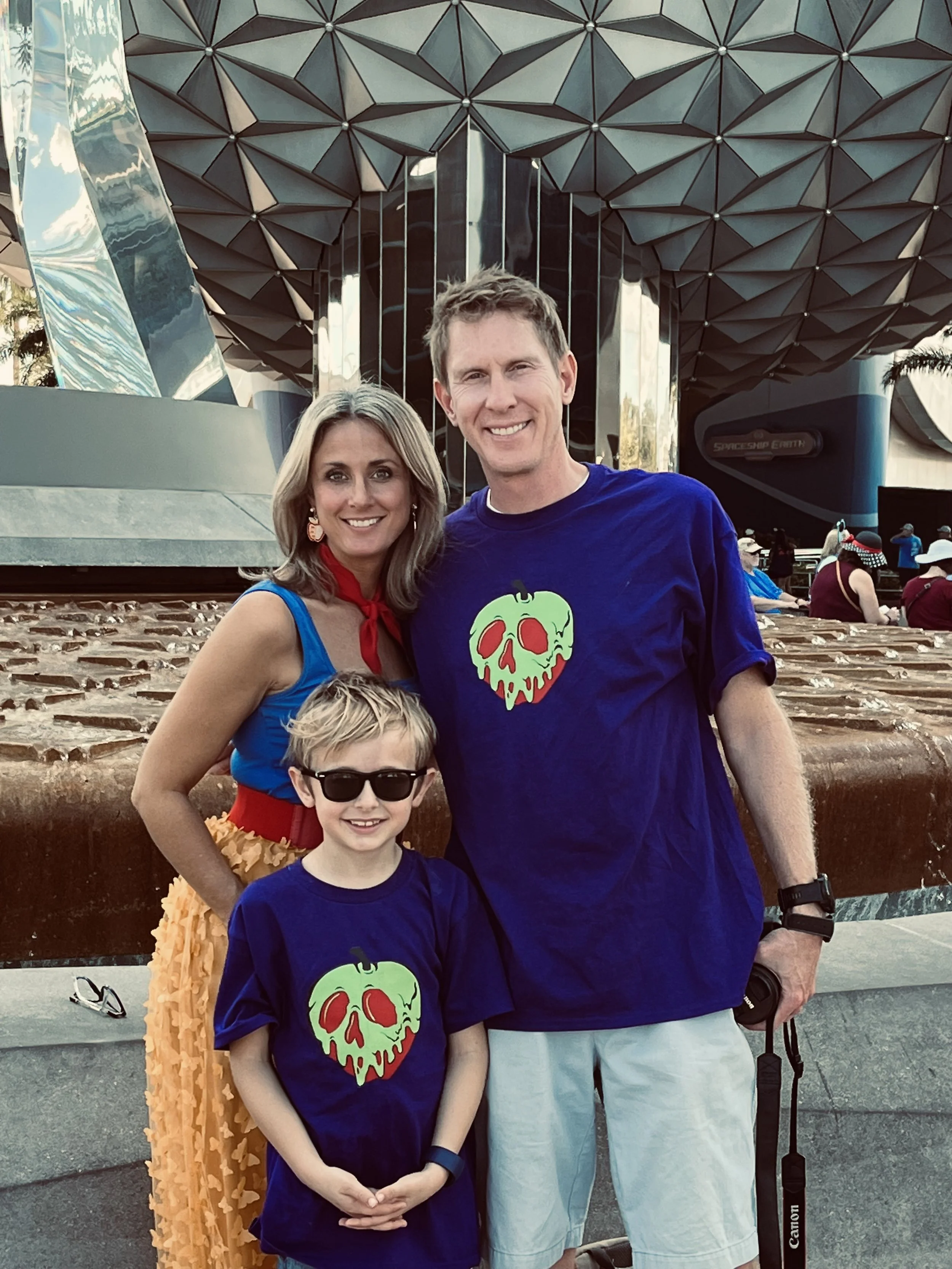 Family of three.  The woman has blonde hair and is wearing a blue top with a red scarf, tand yellow skirt disneybounding as snow white.  husband and son wearing purple shirt with poisonous apples on them