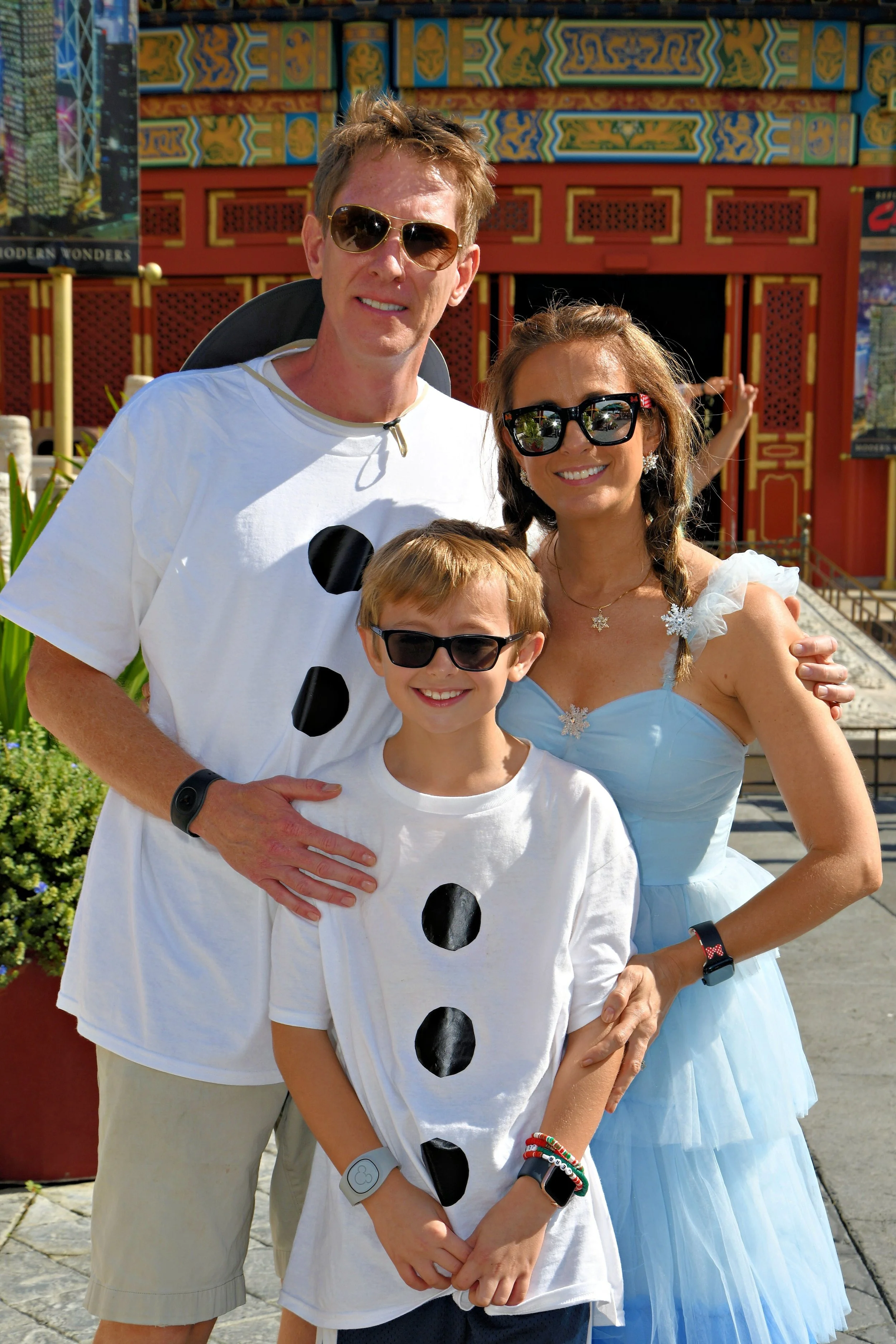 A family of three: a man, a woman, and a boy, smiling and posing together outdoors, with a colorful building in the background.  Woman is disneybounding Elsa and husband and son have on shirts representing olaf.
