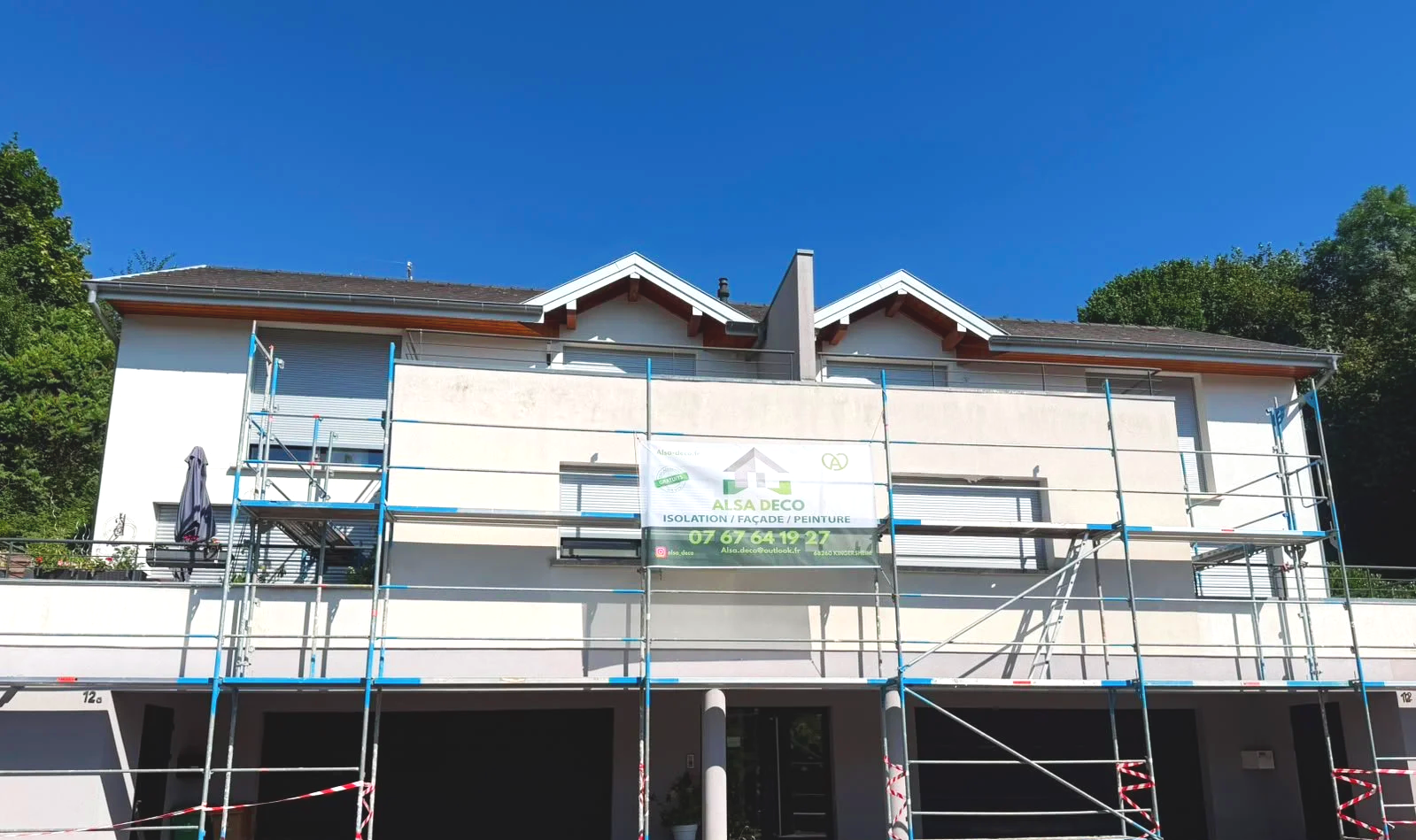 Construction d'un bâtiment résidentiel avec des échafaudages placés devant. Le bâtiment est blanc avec un toit en pente, sous un ciel bleu clair.