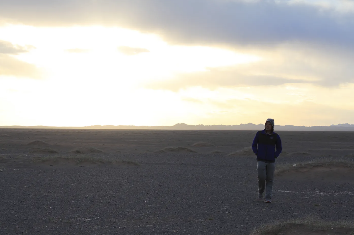 A person in a hoodie walking with their hands in their pockets against the sun setting in the background and mountain ranges