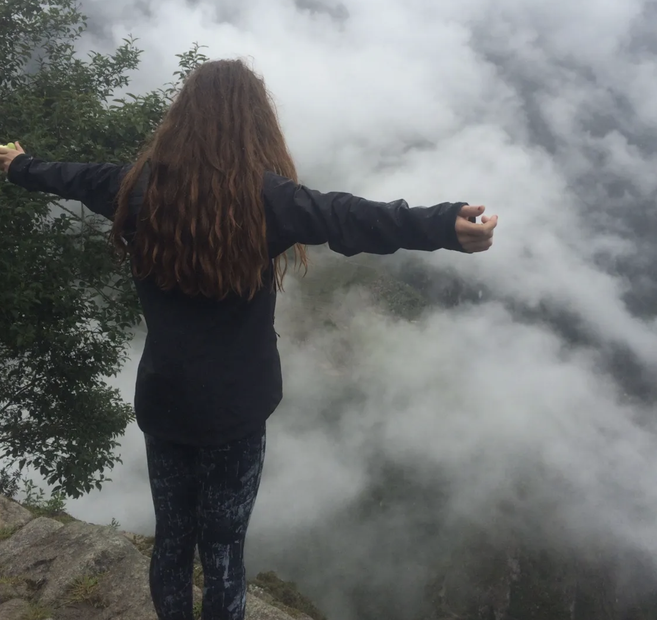A traveling photo of a woman with arms stretched wide standing on a mountain overlooking the clouds