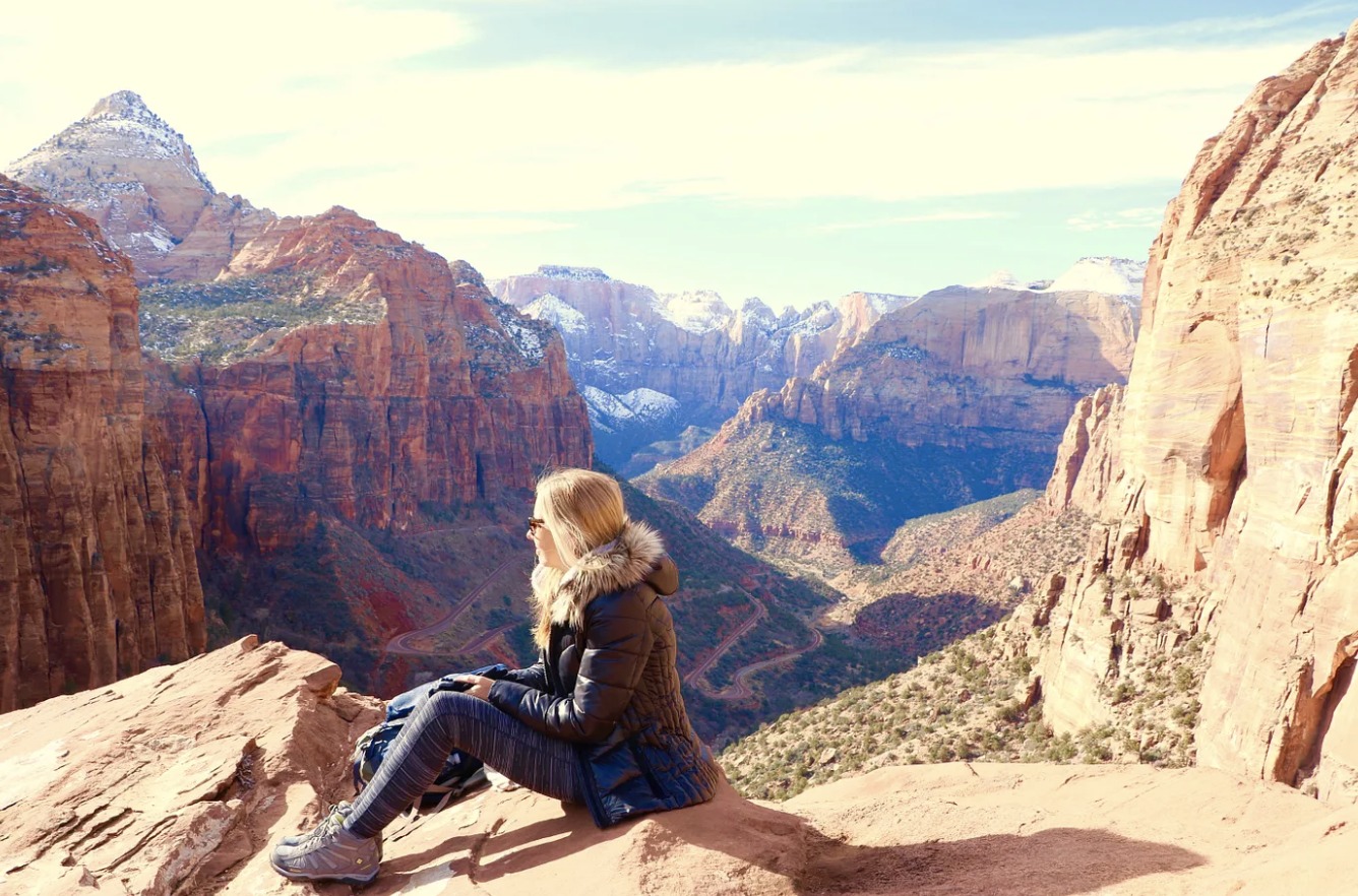 Kirsten Nelson Telan in a coat and hiking boots siting a top of a rock looking over mountains and red rock canyons