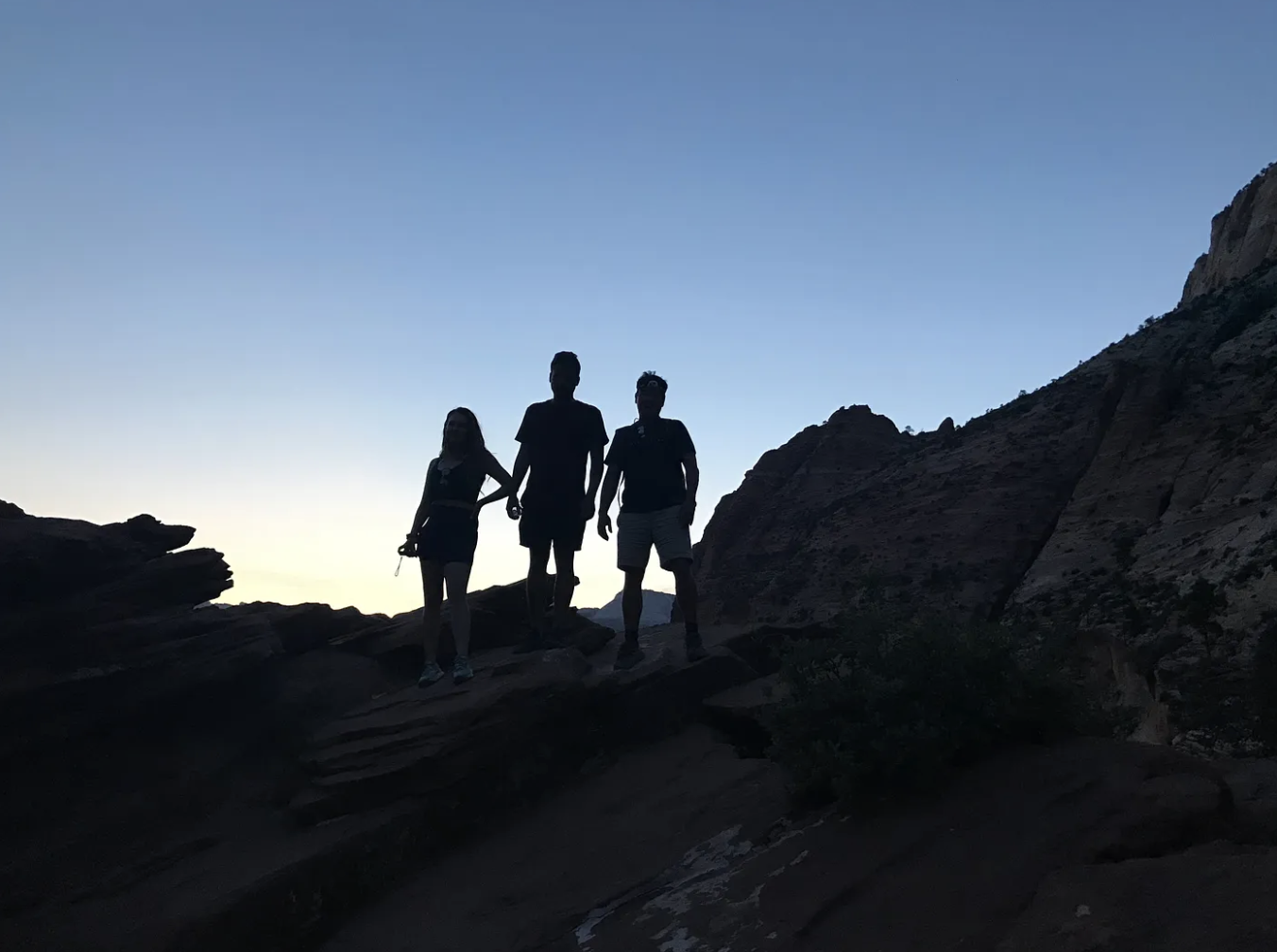 Three silhouettes of people standing on a rocky mountain in Zion National Park with the sun setting in the background