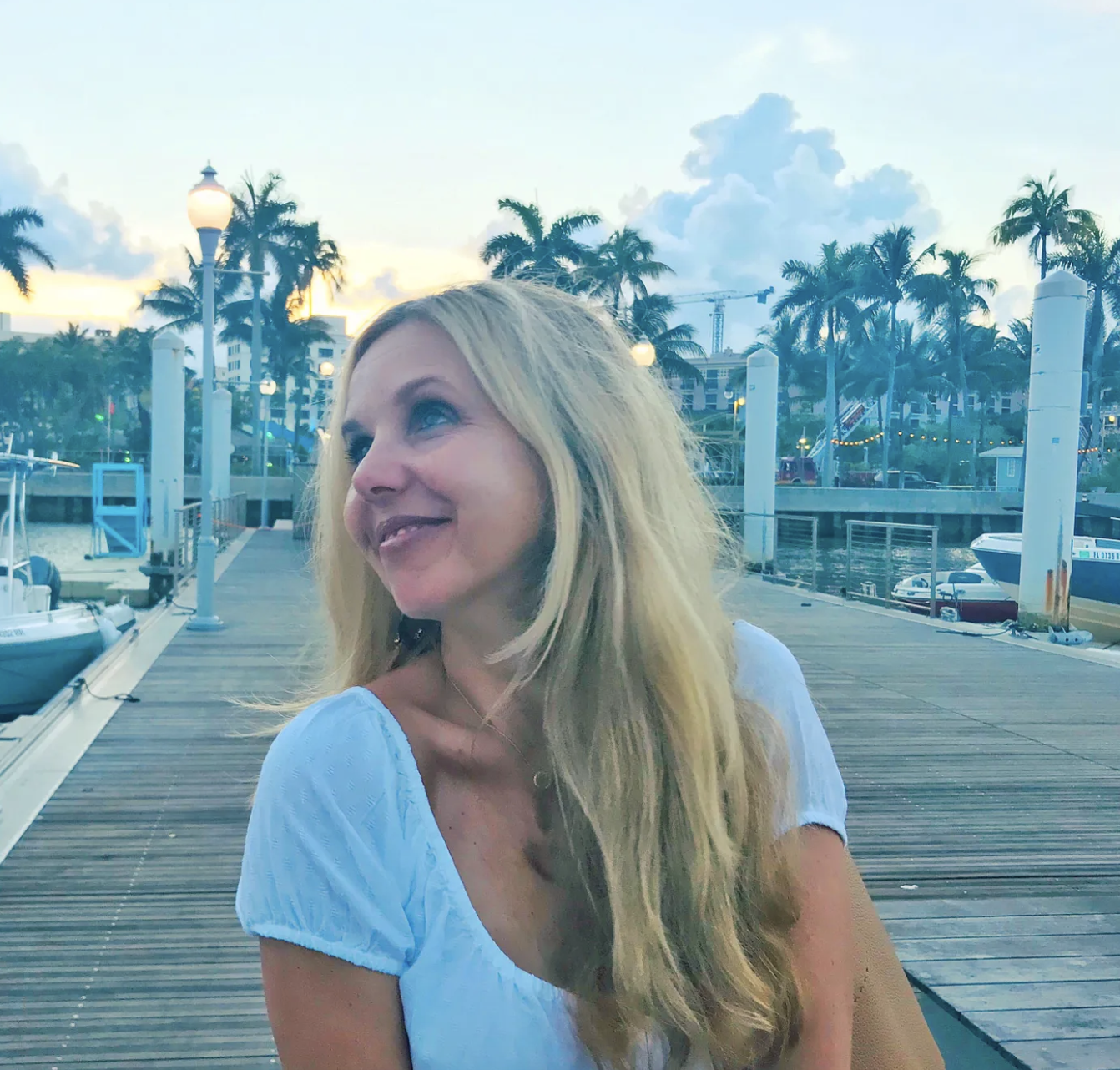 Kirsten Nelson Telan sitting on a boardwalk looking off into the distance smiling with palm trees, boats, water, buildings, and string lights in the background. A vacation photo.