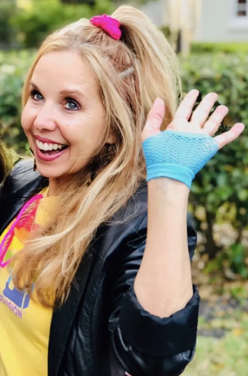 Kirsten Nelson Telan dressed up in an 80s outfit with a scrunchie, gloves, colorful tee shirt and sun glasses,