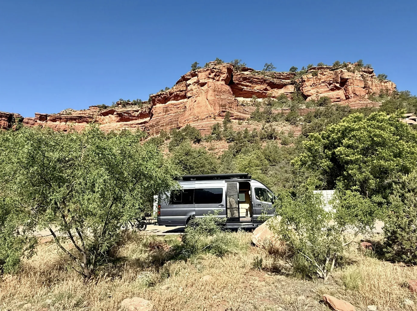 A travel van out west against red rock in the background with grass and trees