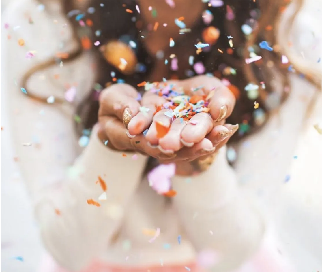 A woman holding a handful of confetti blowing it into the camera so there is a cloud of confetti