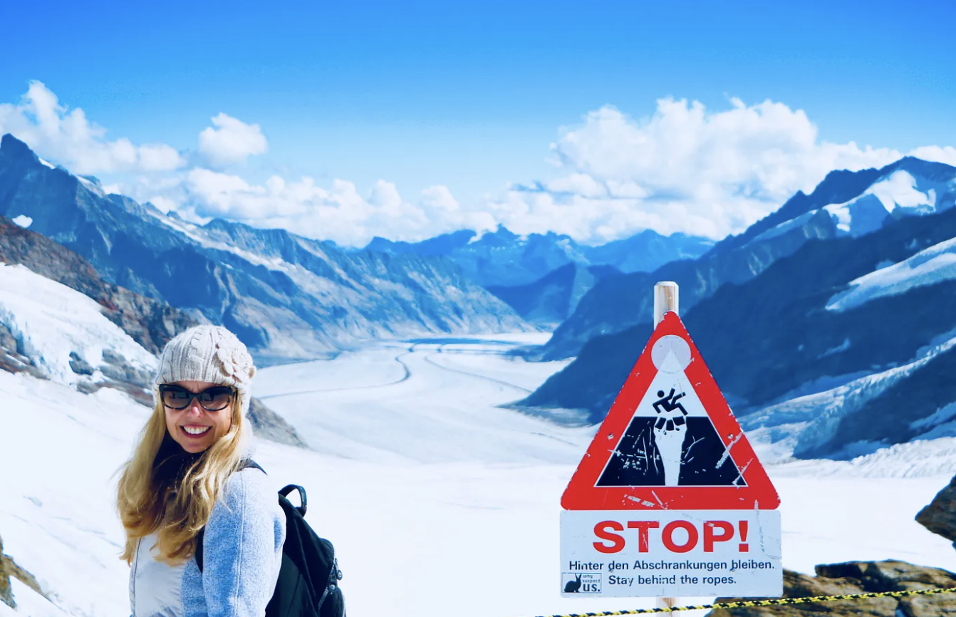 Kirsten smiling in front of a snowy mountain backdrop next to a warning sign that says "Stop stay behind the ropes" in multiple languages