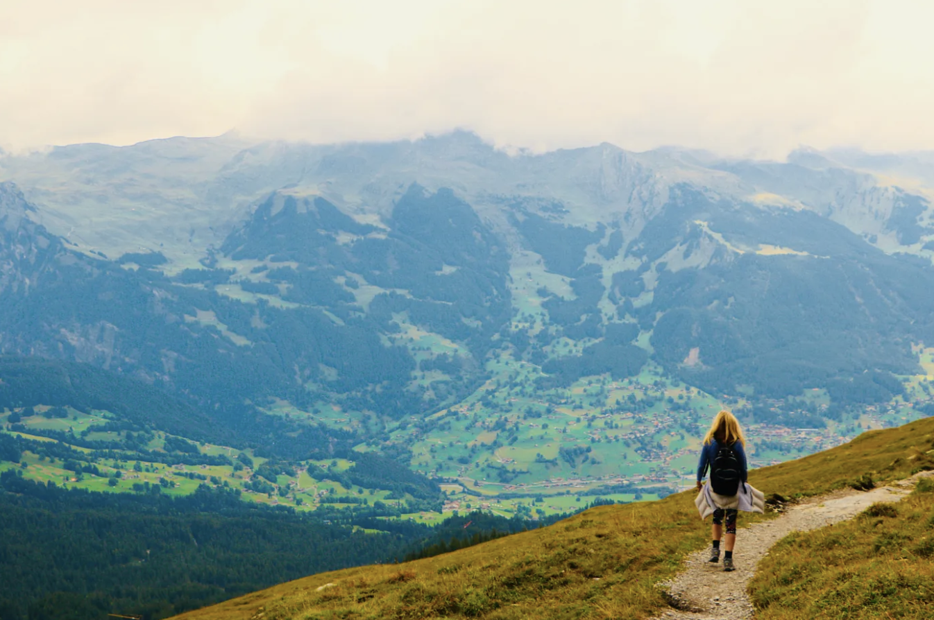 A travel photo of Kirsten Nelson Telan standing on a mountain trail looking out at the vast mountain range