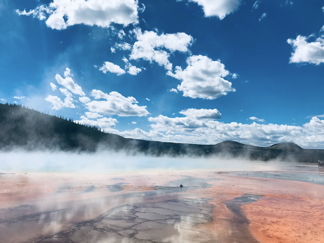 Blue sunny sky with fluffy while clouds over a mountain range and mist rising from red sand and water