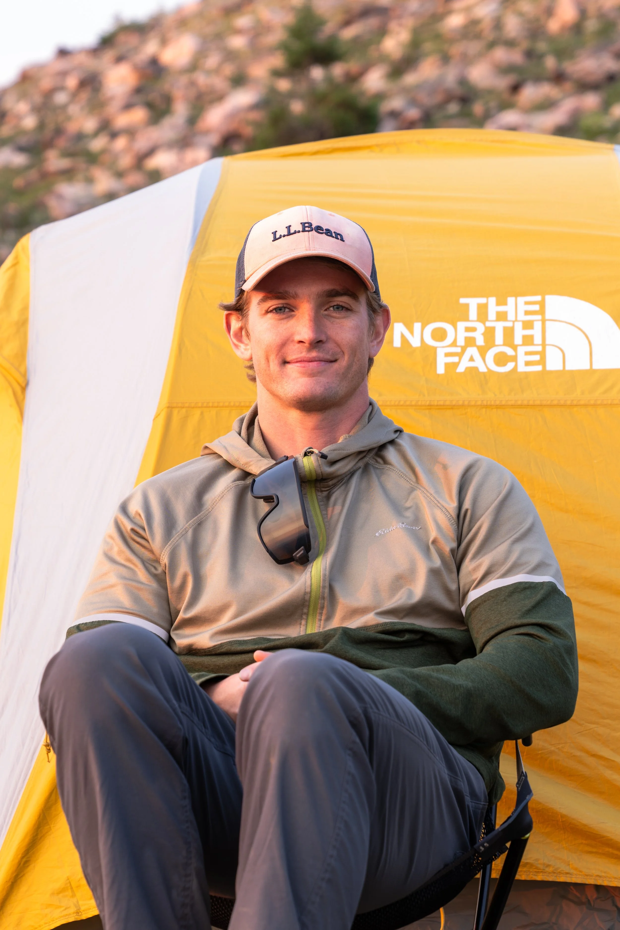 A smiling young man seated by a yellow and white tent bearing The North Face logo, wearing a beige and green Patagonia jacket, a pink and beige L.L. Bean cap, and black goggles hanging around his neck, outdoors with a rocky hillside in the background.