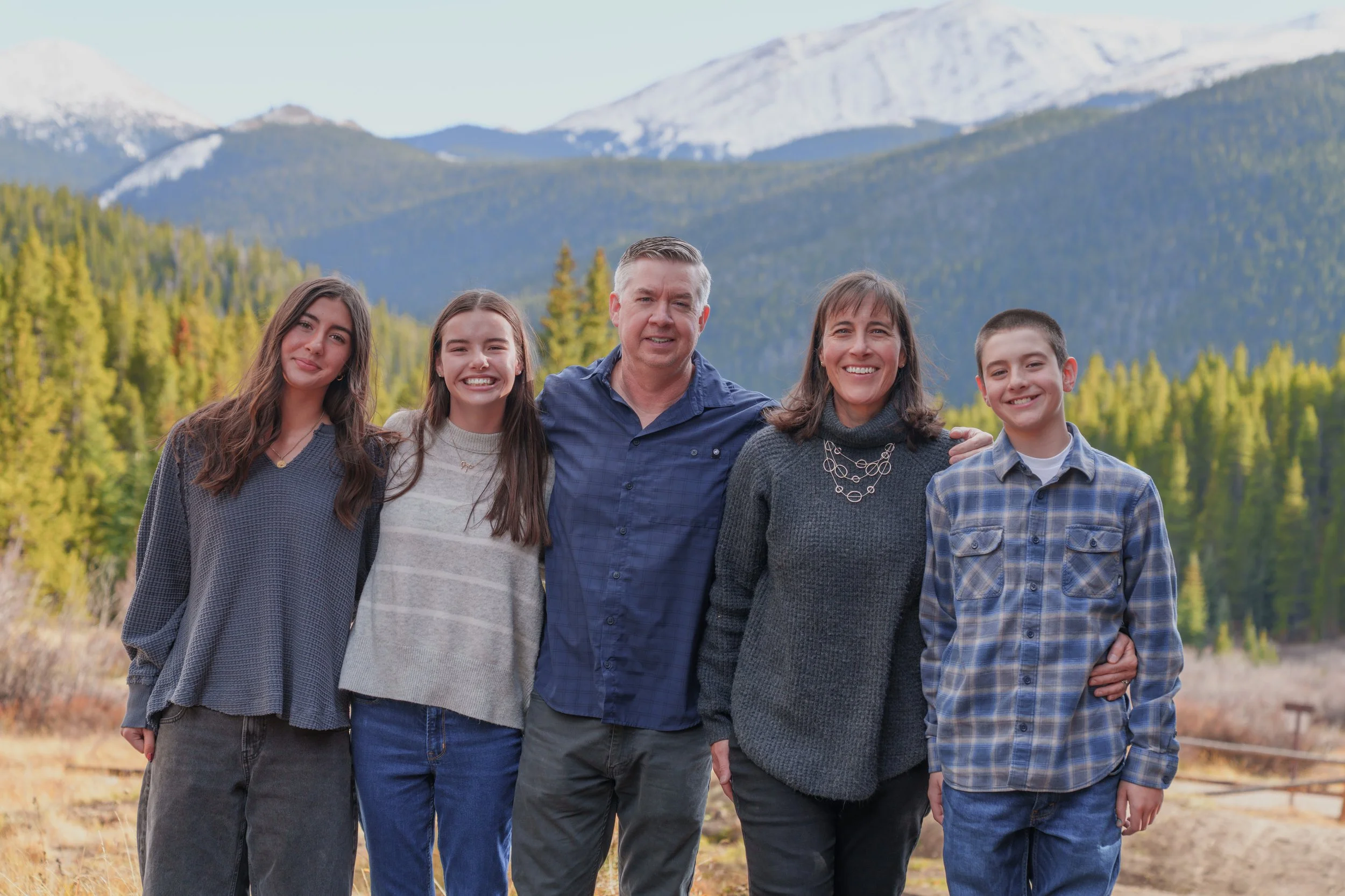 Family of five standing outdoors in front of mountains and forest, smiling at the camera.