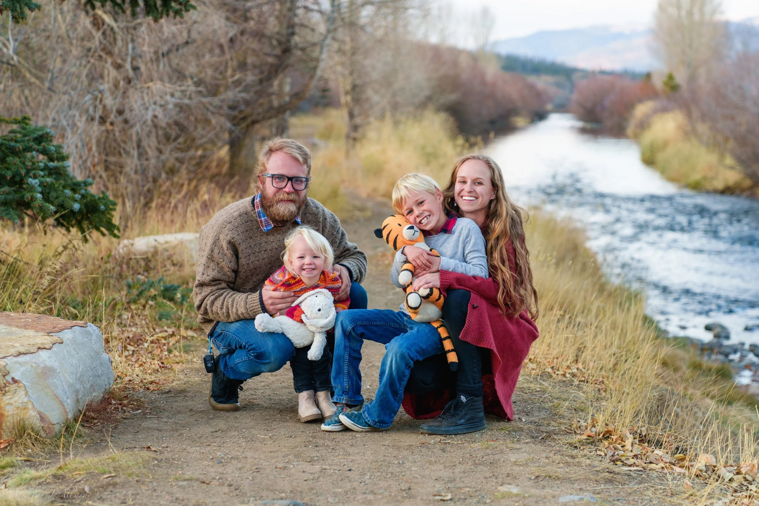 Family of four posing outdoors on a path by a river during fall, with trees and mountains in the background. The father, mother, and two children are smiling and holding stuffed animals.