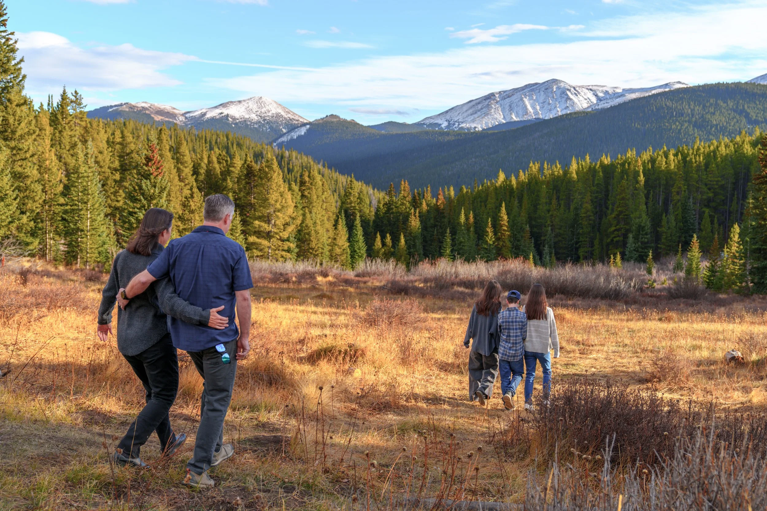 A group of five people hiking in a mountain landscape with evergreen trees and snow-capped peaks in the background.