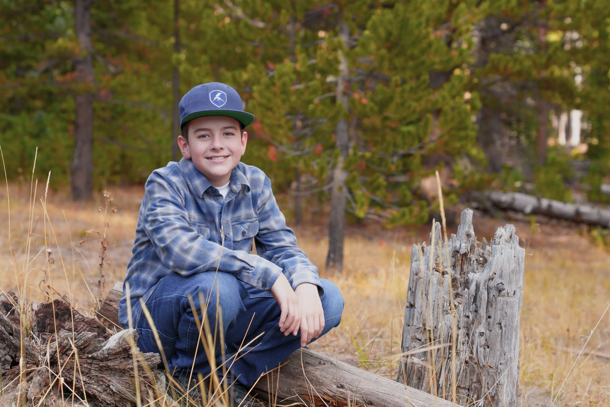A young boy in a plaid shirt and baseball cap sitting on a fallen log in a forested area with trees and dry grass.