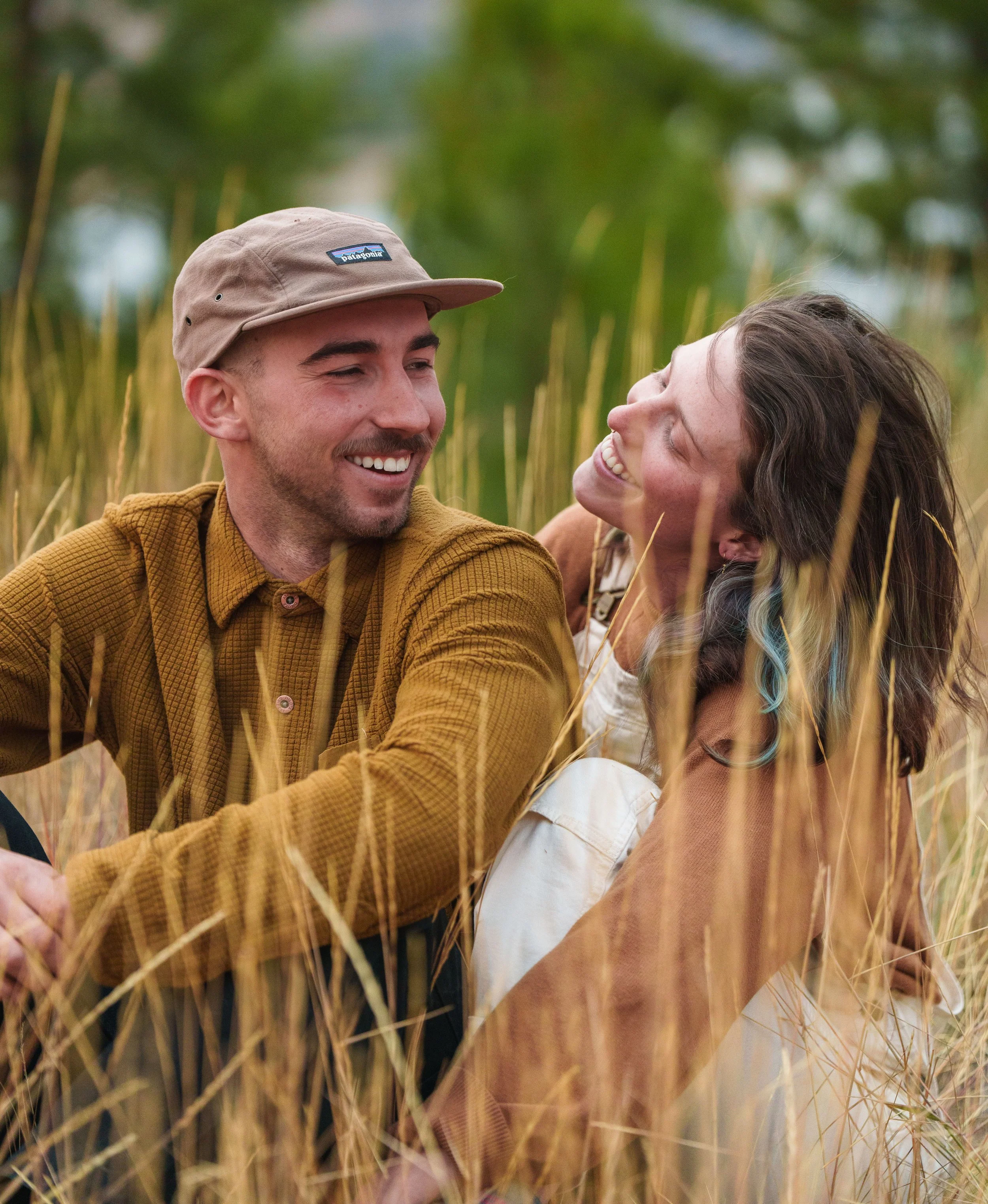 Two people smiling and sitting in a field of tall grass, enjoying each other's company.