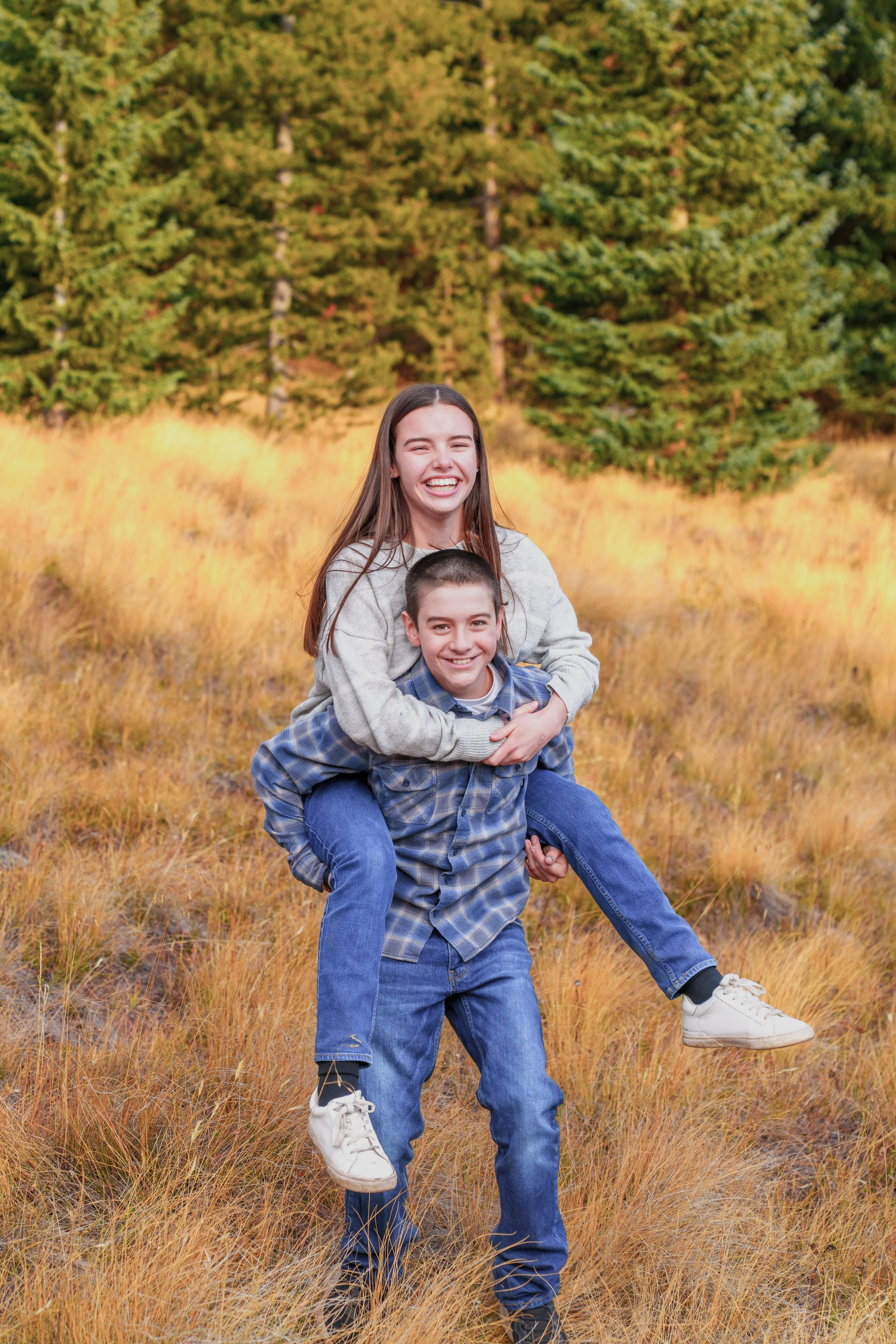 A young girl and boy laughing and playing outdoors in a grassy field with trees in the background. The girl is on the boy's back, and they appear to be happy.