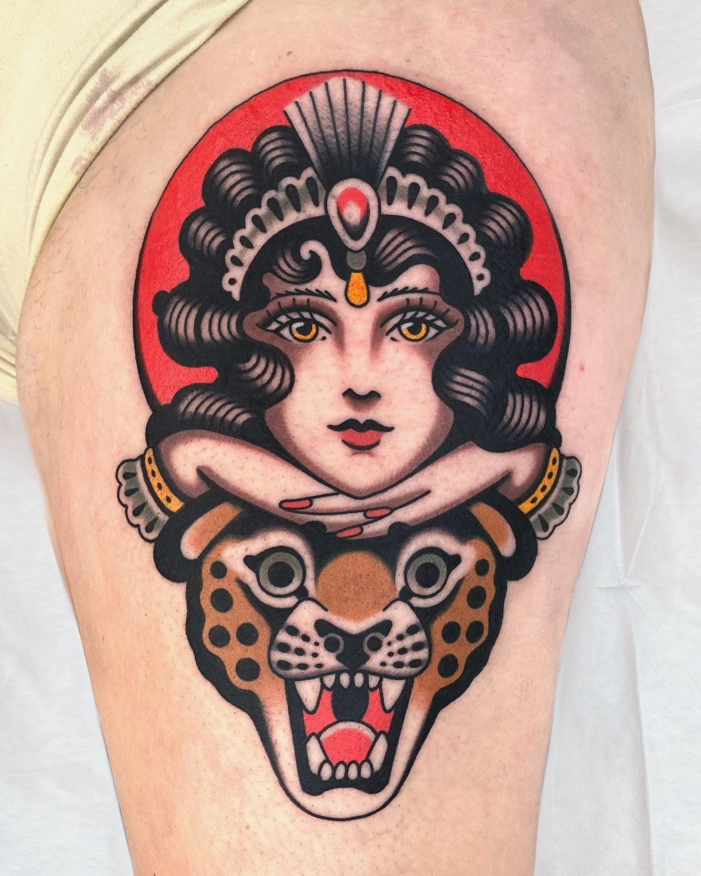 Tattoo of a woman's face with long black curly hair, crowned with jewelry, resting her chin on her hands above a snarling jaguar's head, in traditional style with bold black outlines and vibrant colors.