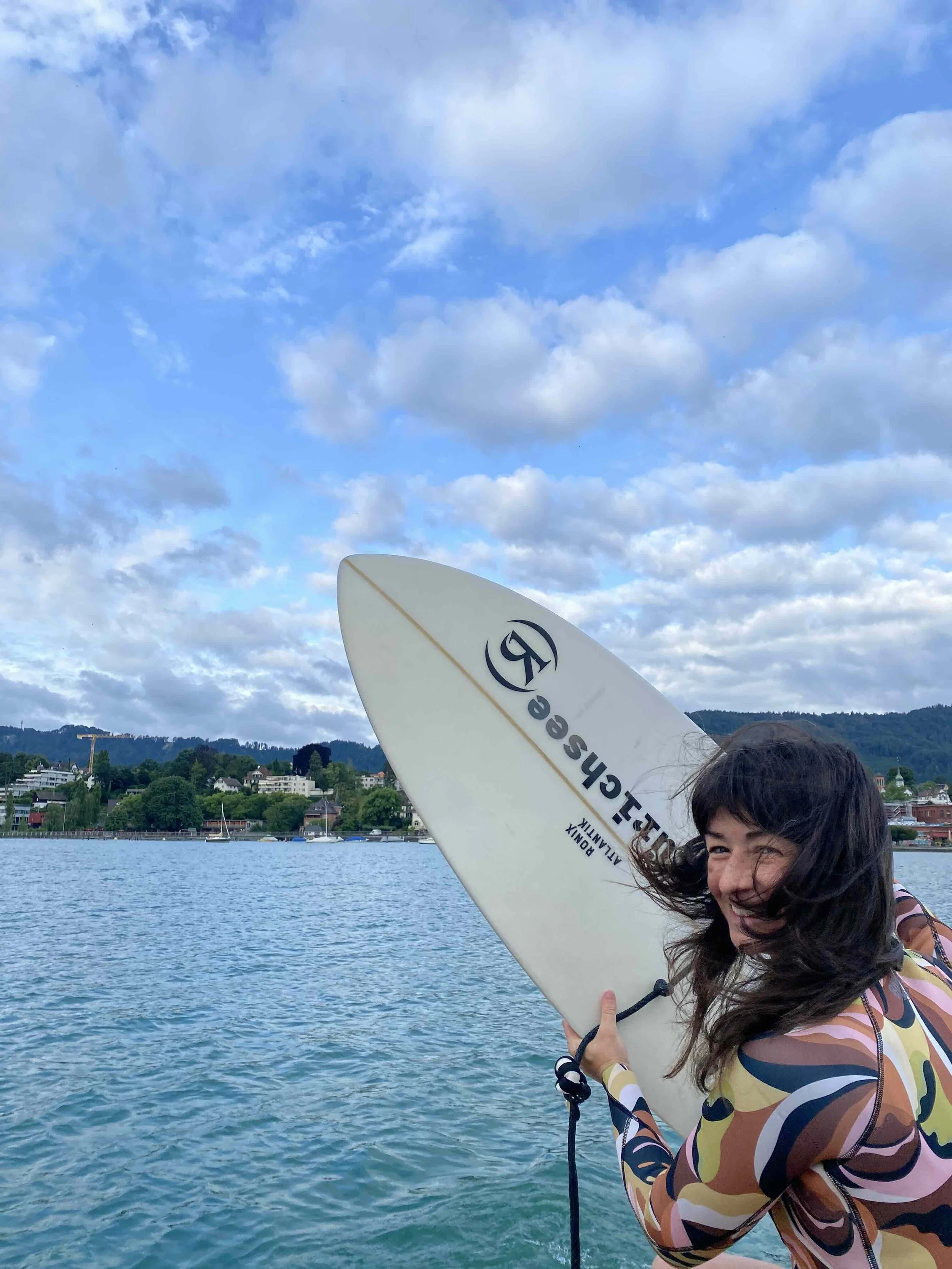 Frau hält ein Surfbrett am Wasser, lächelnd und im Wind, mit einer ländlichen Gegend im Hintergrund.