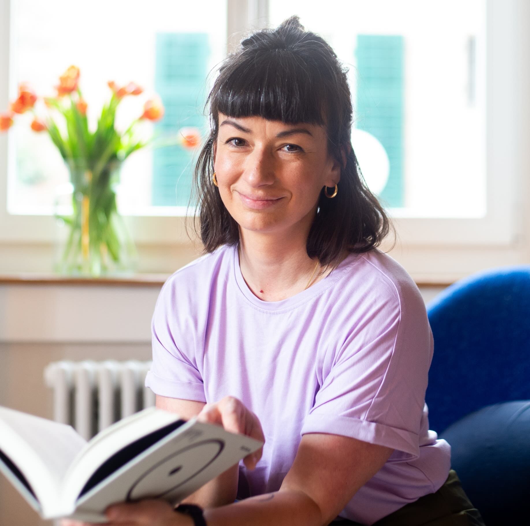 Eine lächelnde Frau mit dunklem Haar und kurzen Pony, trägt eine lavendelfarbene T-Shirt, sitzt in einem hellen Raum mit blauen Sessel, hält ein Buch mit geometrischem Muster in den Händen, im Hintergrund eine Vase mit orangefarbenen Blumen auf einem Fensterbrett.