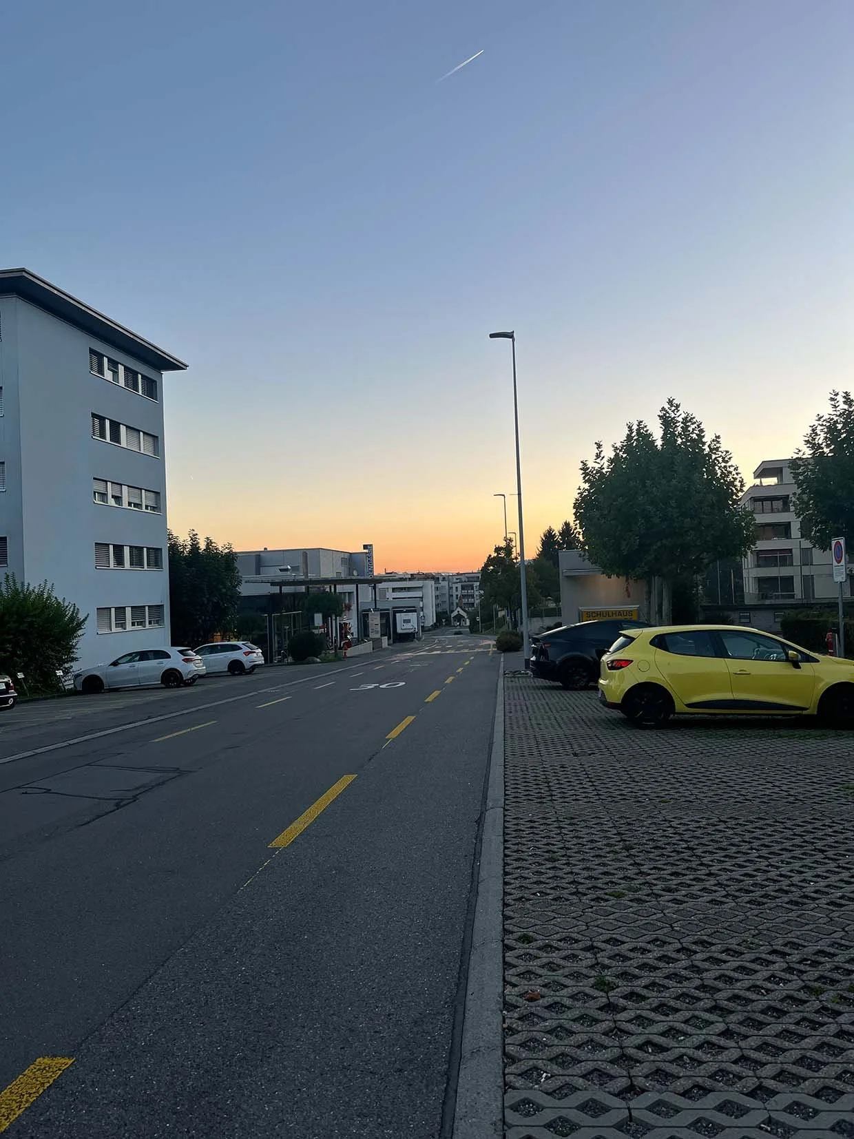 Straßenansicht bei Sonnenuntergang mit Parkplätzen, mehrstöckigen Gebäuden und Bäumen