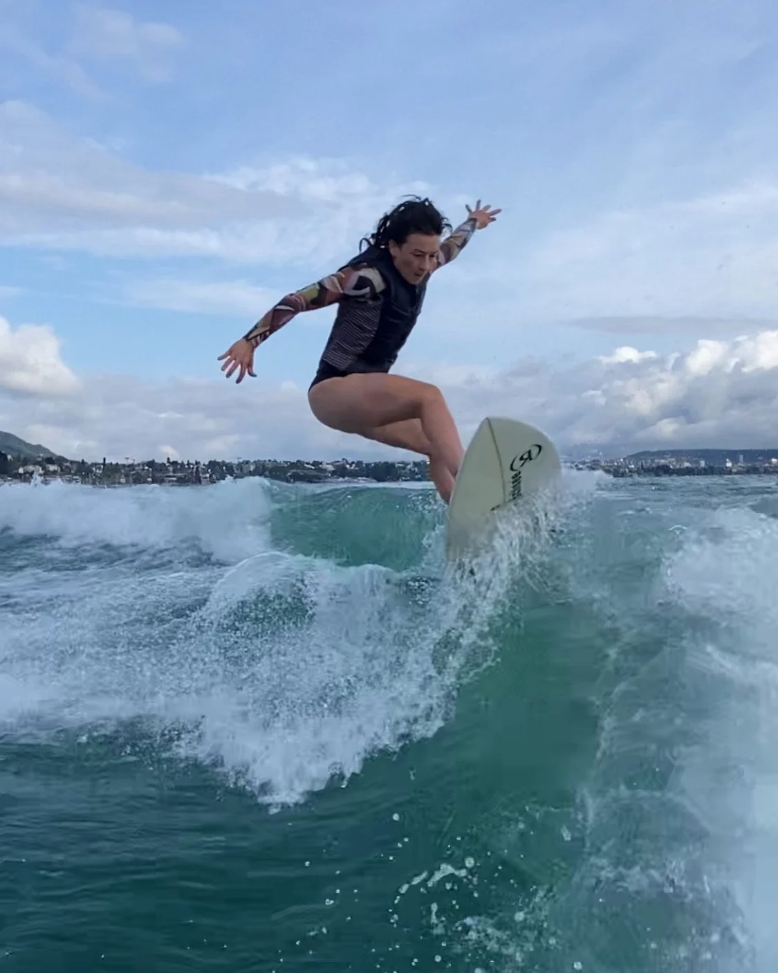 Frau beim Surfen auf einer Welle im Wasser, mit Stadt im Hintergrund und blauem Himmel.