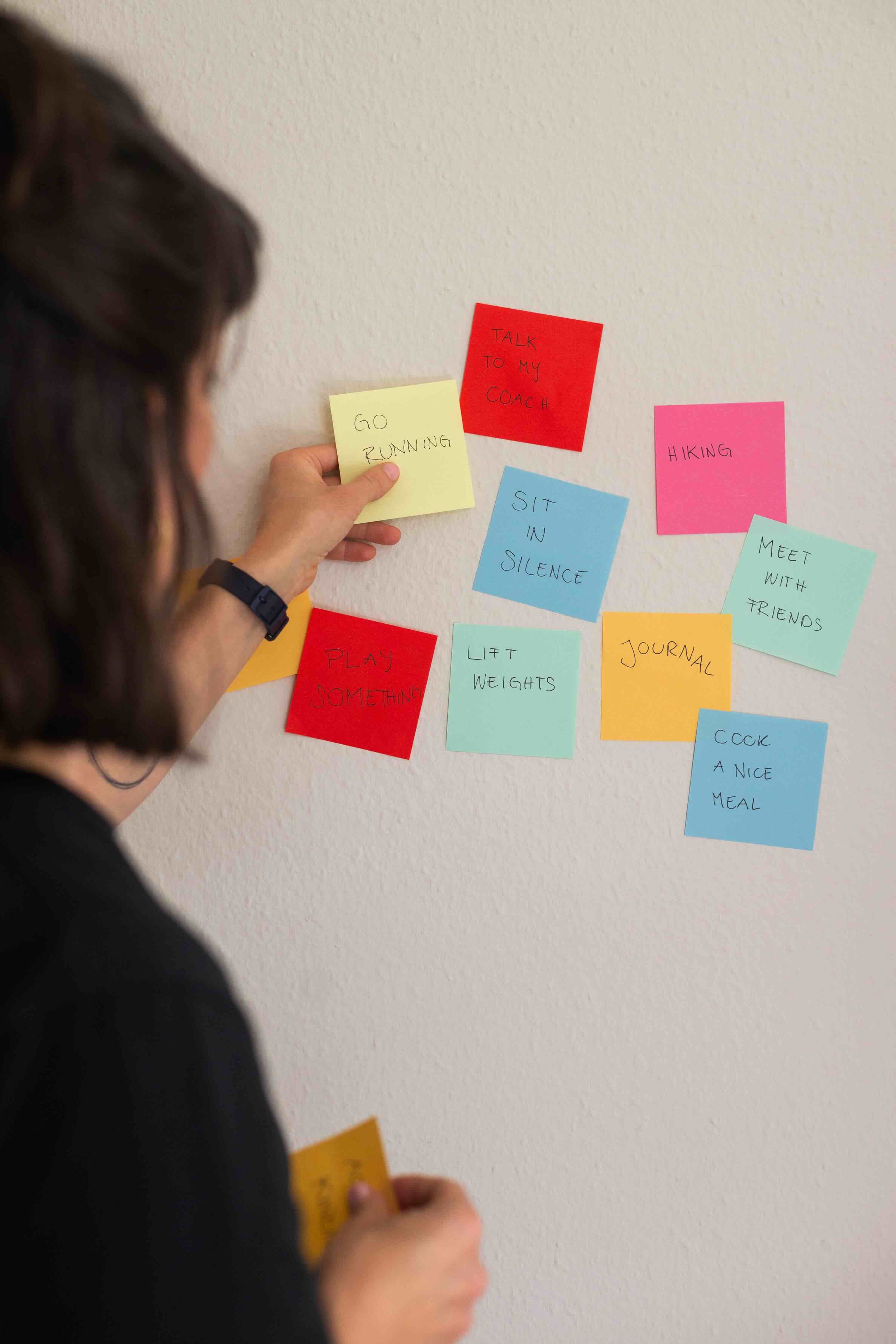 Frau hängt bunte Haftnotizen an die Wand mit Aktivitäten wie 'Laut sprechen', 'Spazieren gehen' und 'Kochen'.