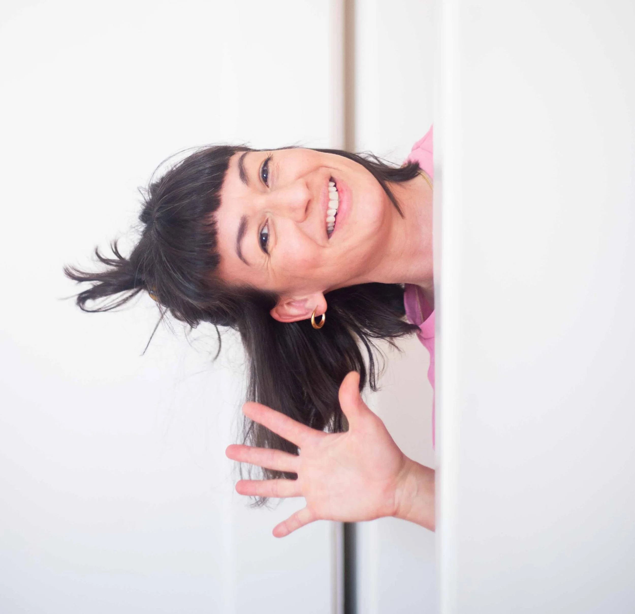 Lächelnde Frau mit dunklen Haaren und Ohrringen, winkend, im pinkfarbenen Top, im Büro oder zu Hause.