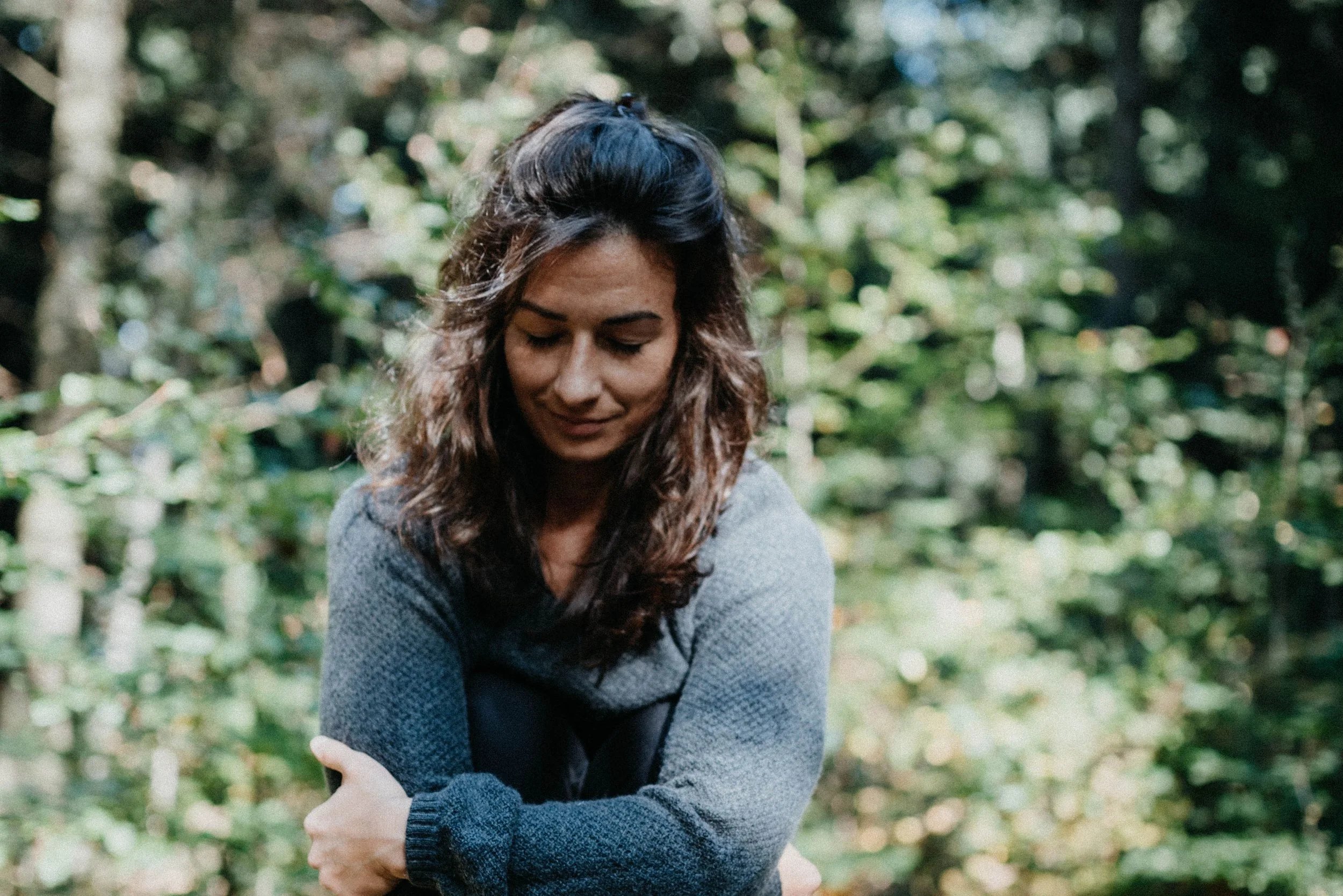 Frau mit braunen Locken umarmt sich selbst im Wald, trägt grauen Pullover, sonniges Licht in der Natur.