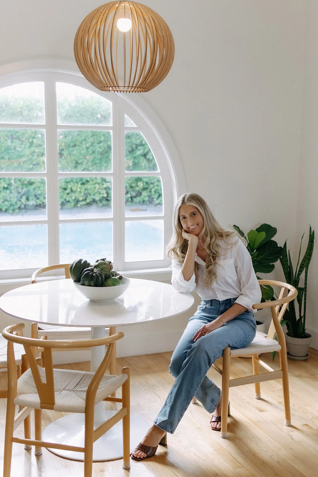 A woman with long blonde hair sitting at a round white table in a bright room with a large arched window, a bowl of squash on the table, and a potted plant nearby, smiling at the camera.