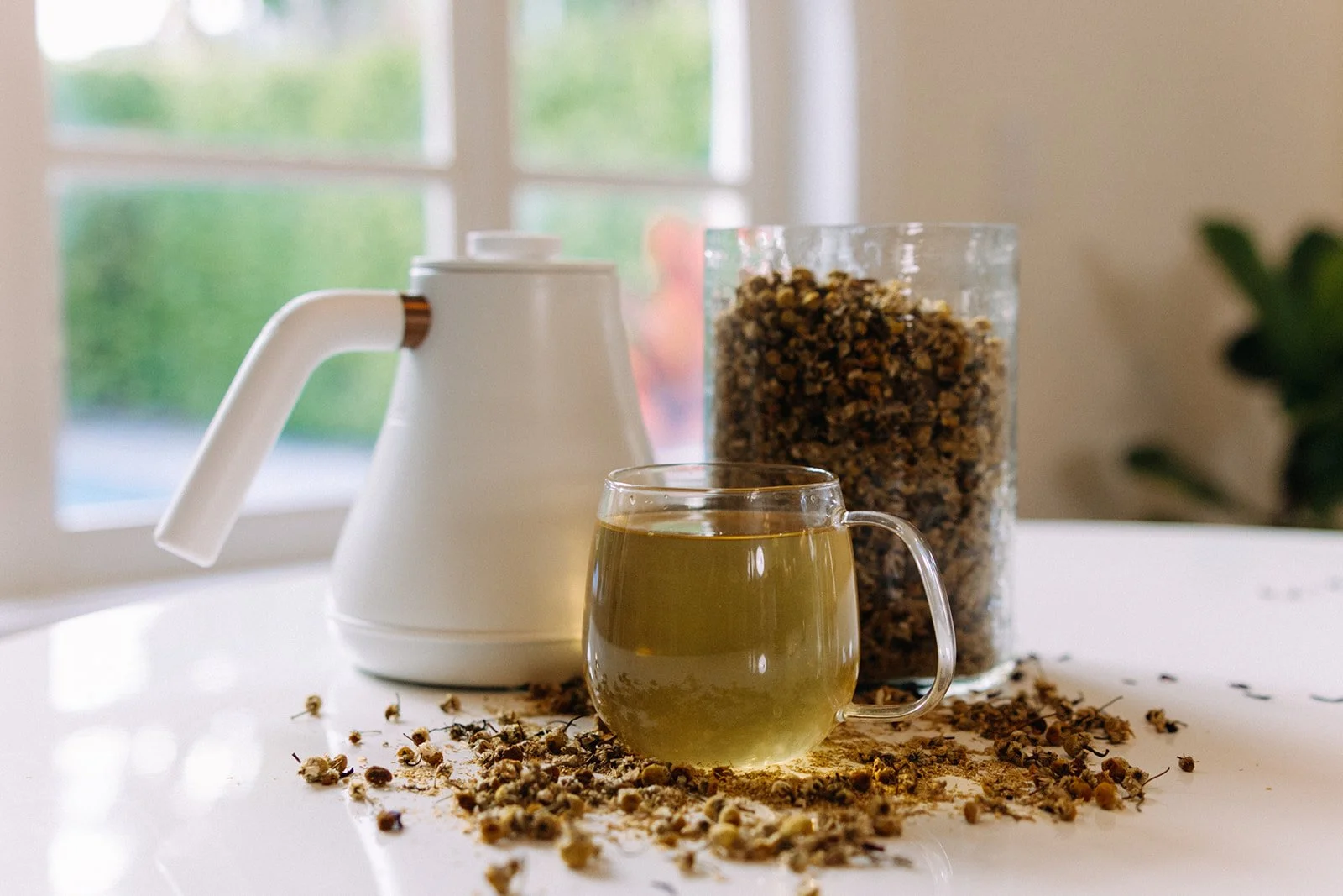 A glass mug of chamomile tea, a white teapot, and a jar of dried chamomile flowers on a white surface near a window.