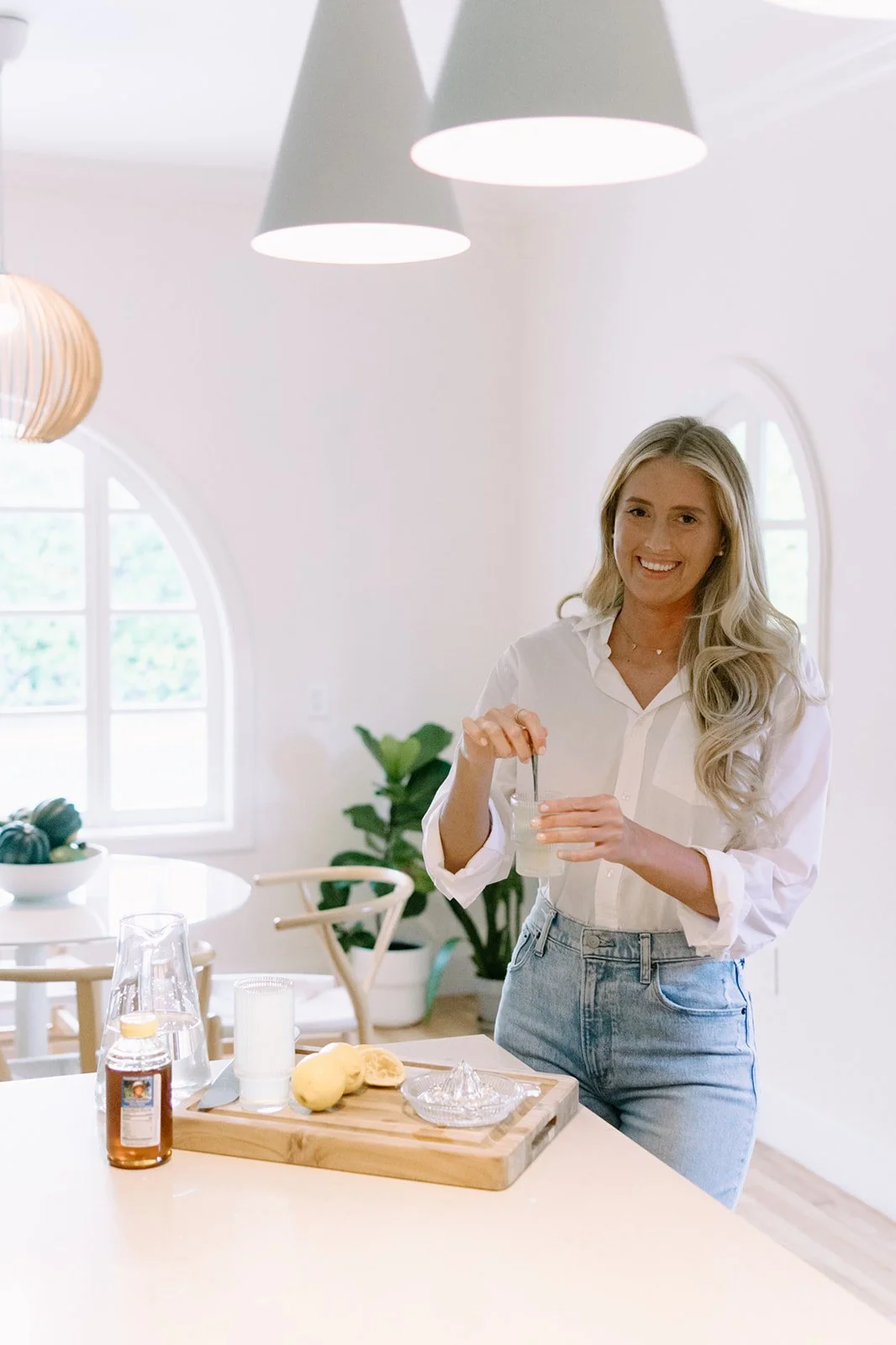 A woman with long blonde hair in a white blouse and jeans is smiling and preparing a drink in a bright, modern kitchen with white walls, arched windows, and Scandinavian-style decor.