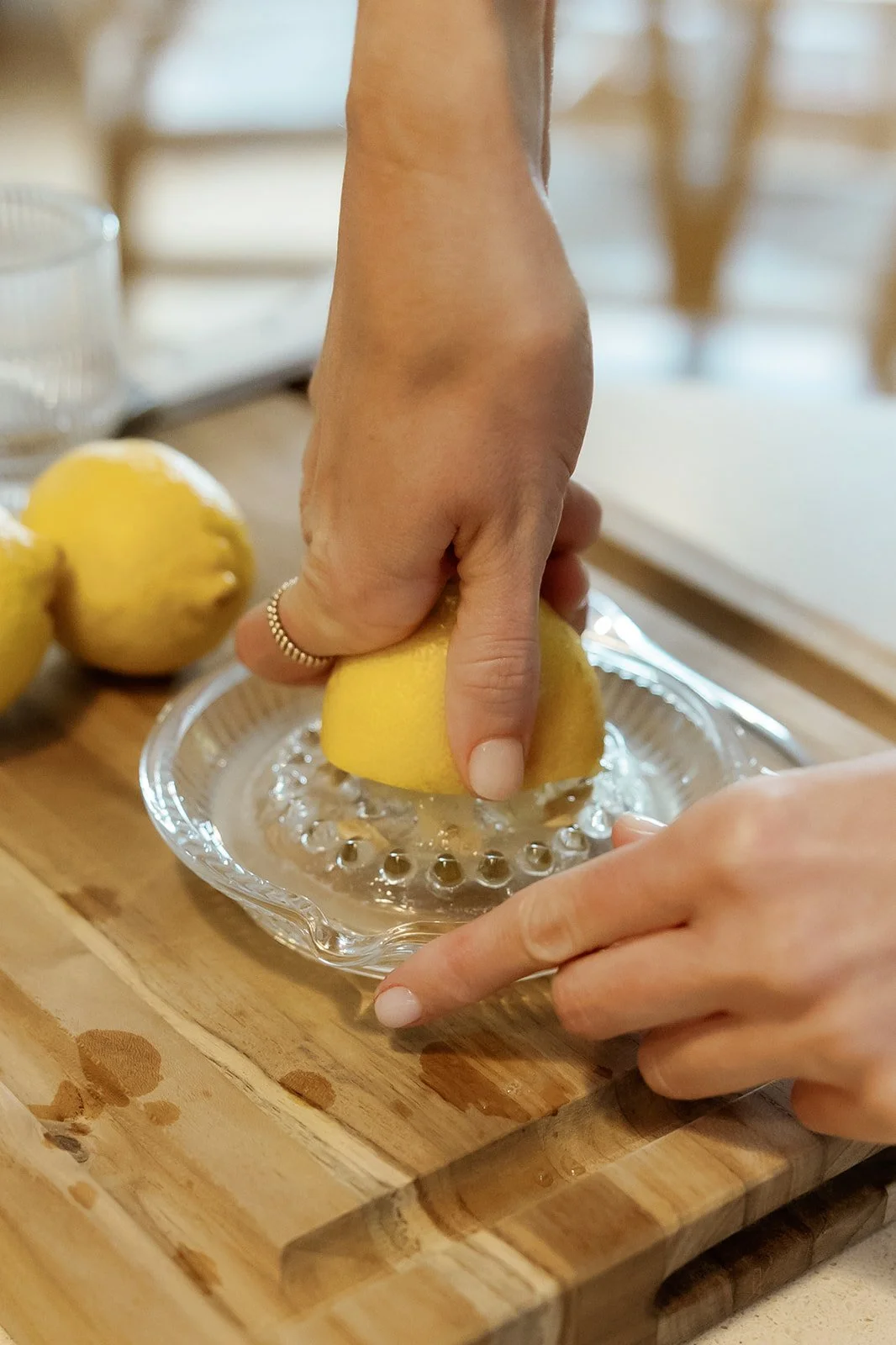 Person squeezing a lemon over a glass dish on a wooden cutting board, with additional lemons in the background.