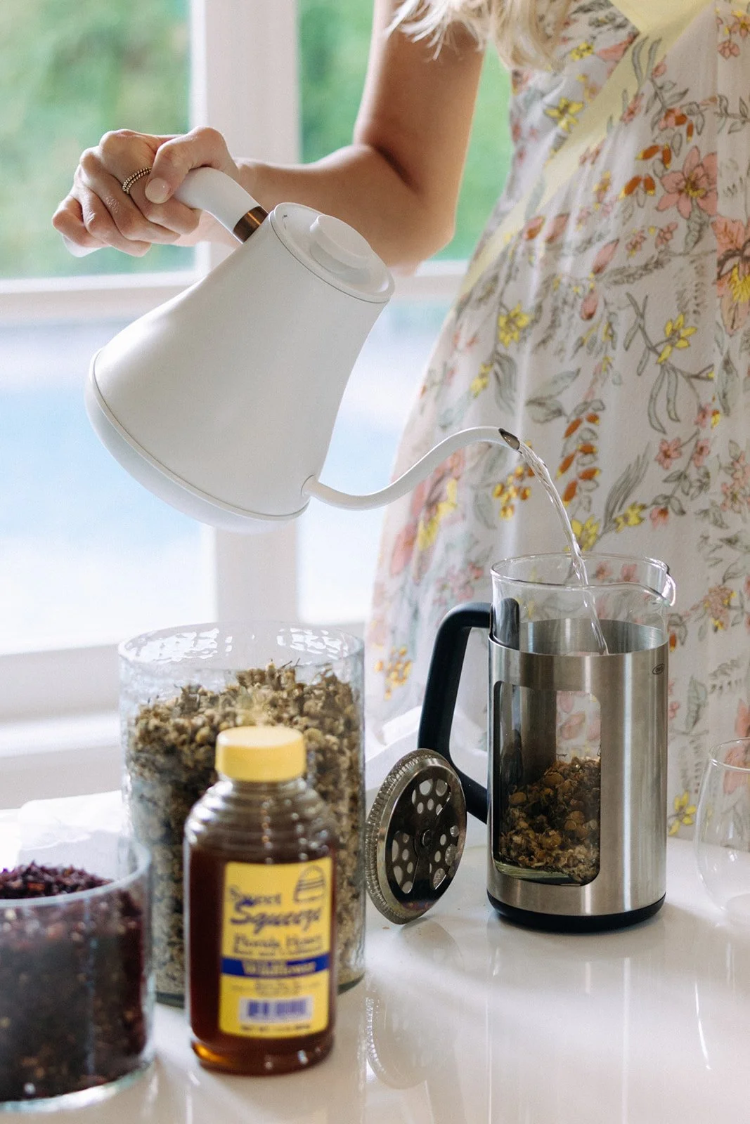 A woman using an electric tea kettle to pour hot water into a glass mug, with various jars of herbs and spices on the kitchen countertop.