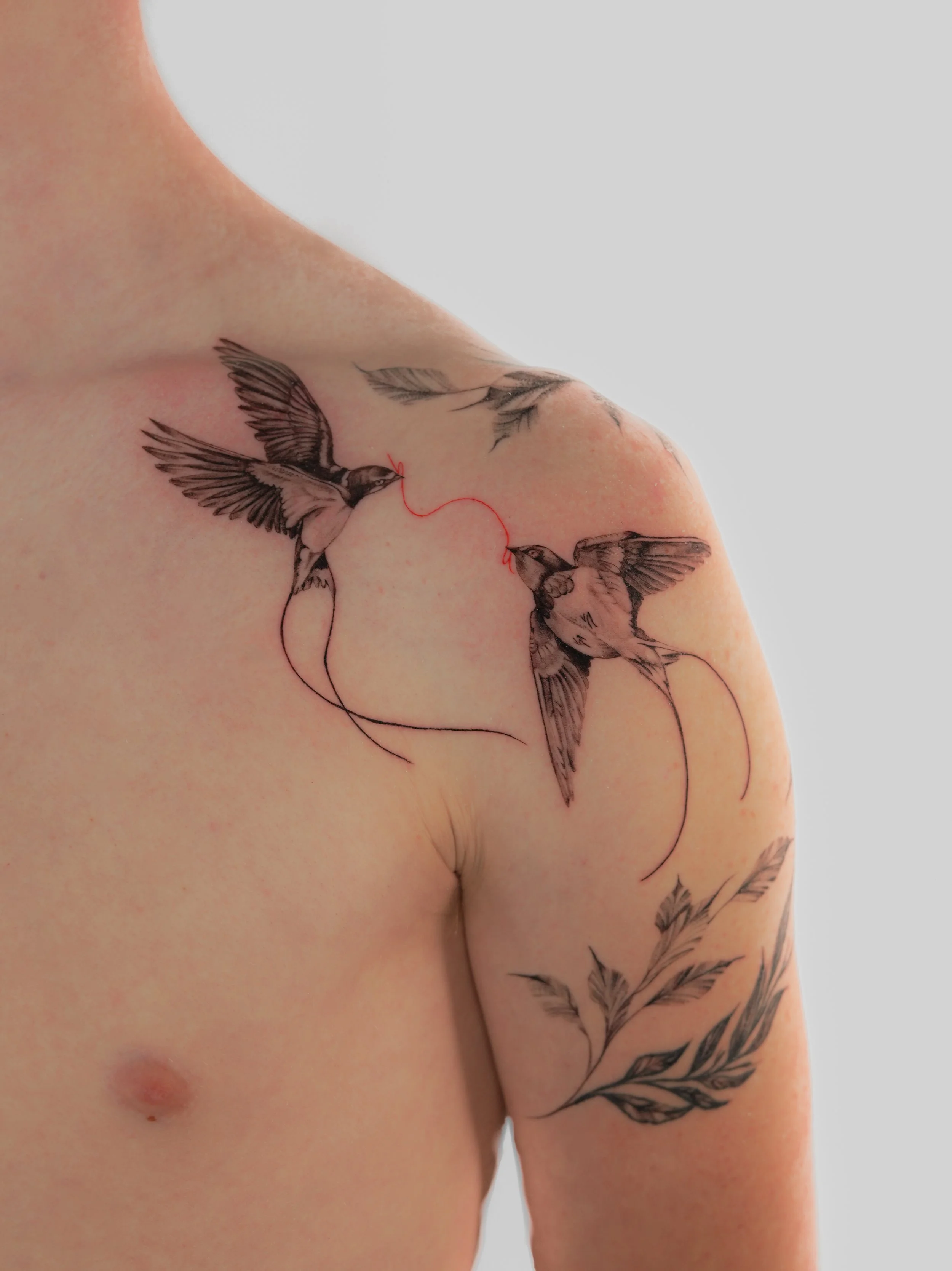 Tattoo of two swallows connected by red thread on person's shoulder combined with delicate floral piece. Done by famous tattoo artist located in Financial District of New York City - Gregory Mirzoyan @gmtattooing