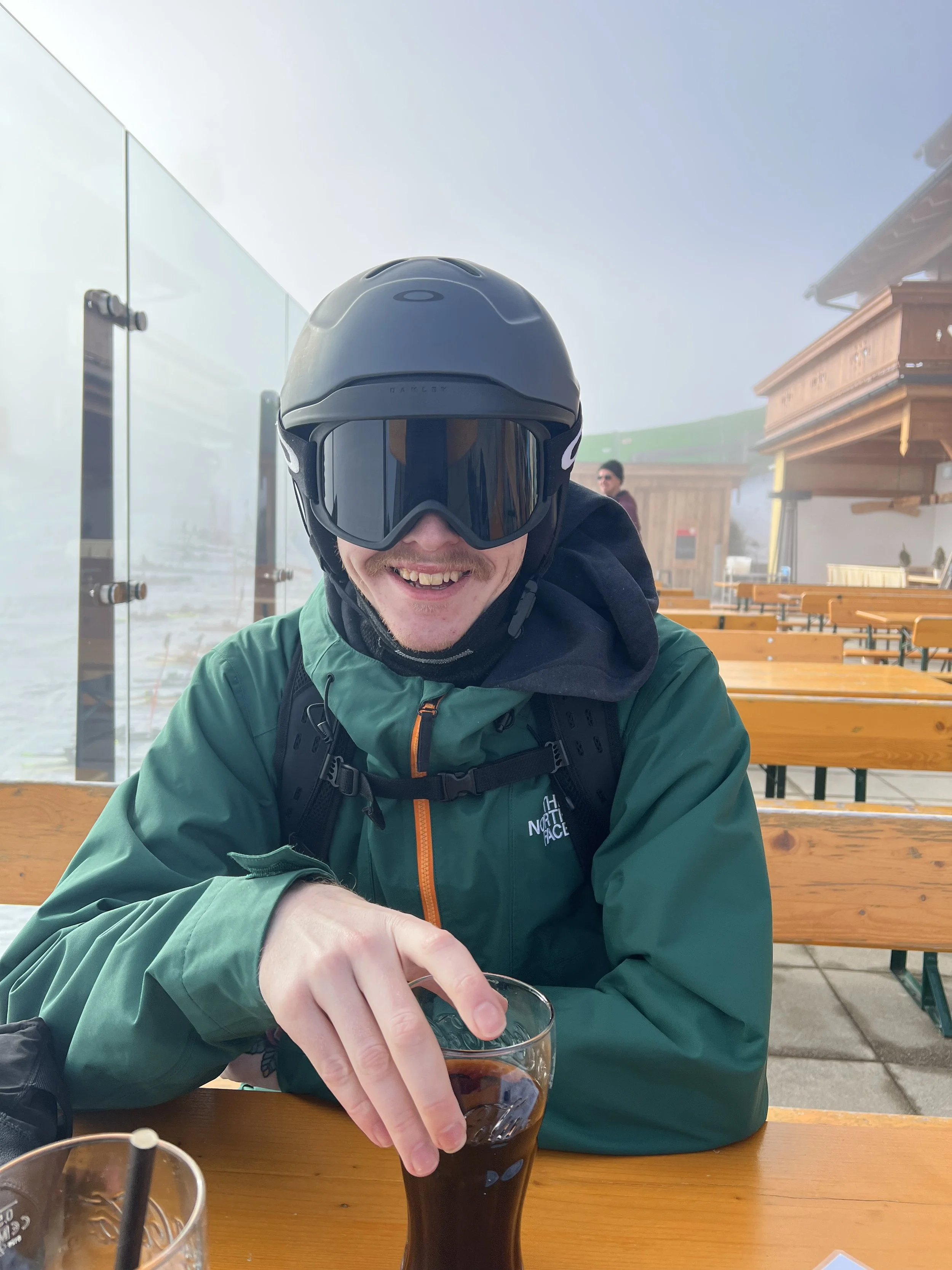 A man wearing a helmet and ski goggles smiling while holding a glass of soda at an outdoor seating area on a foggy day.
