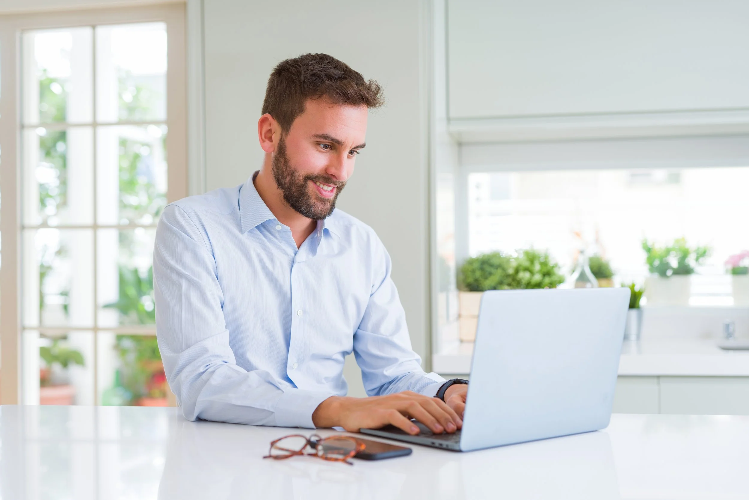 Man with glasses on a white table working on a laptop in a bright kitchen.