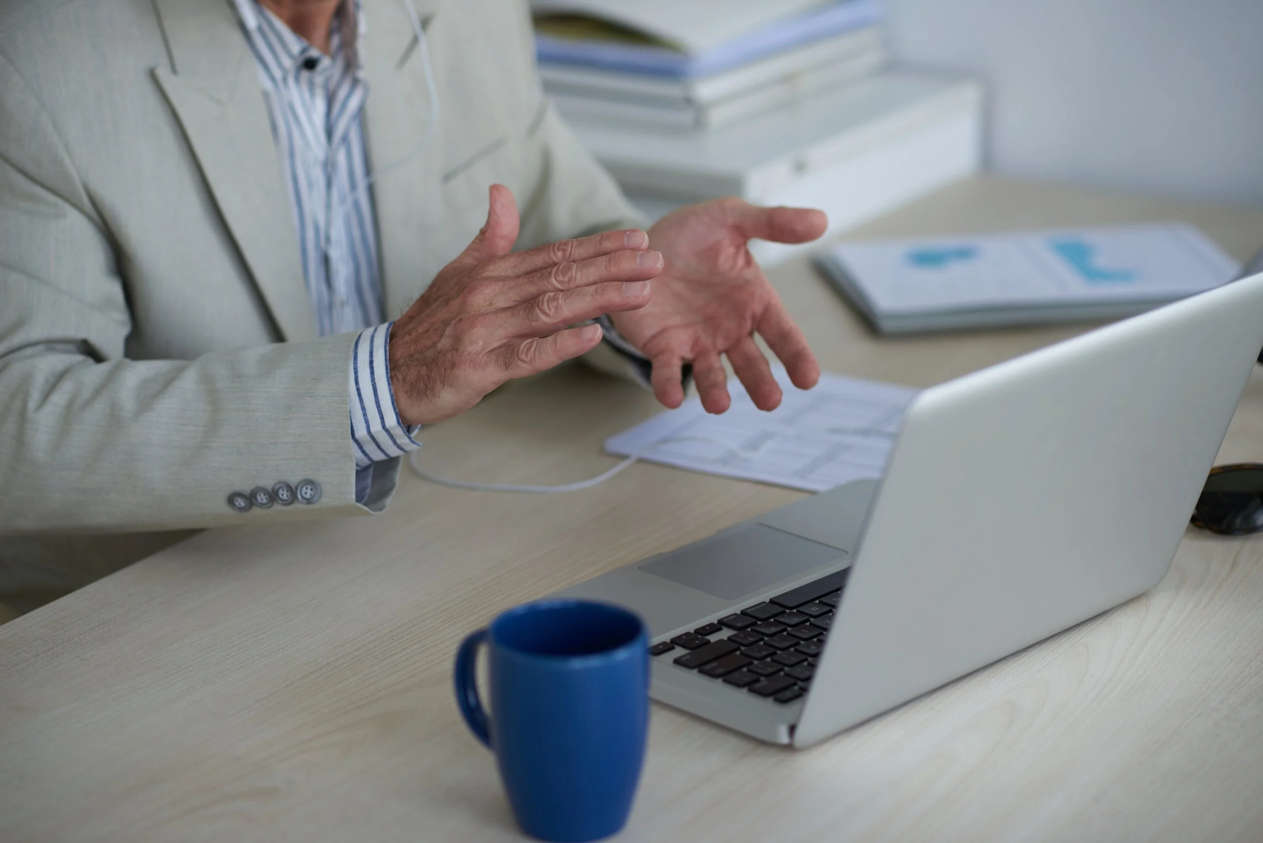 Person in a beige blazer and striped shirt gesturing with hands in front of a laptop on a desk with a blue mug, documents, and stacks of papers.