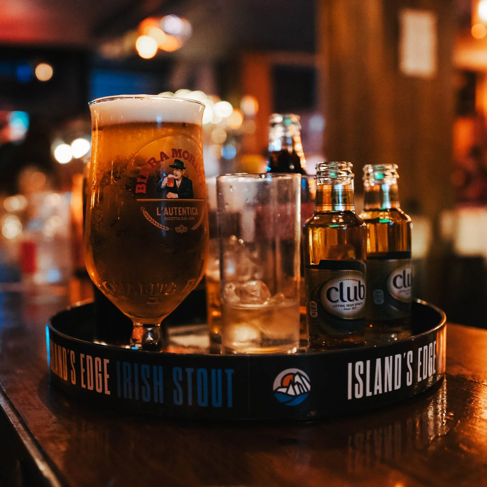 A tray with a glass of beer, several small bottles of club soda, and a glass with ice on a wooden table in a bar setting.
