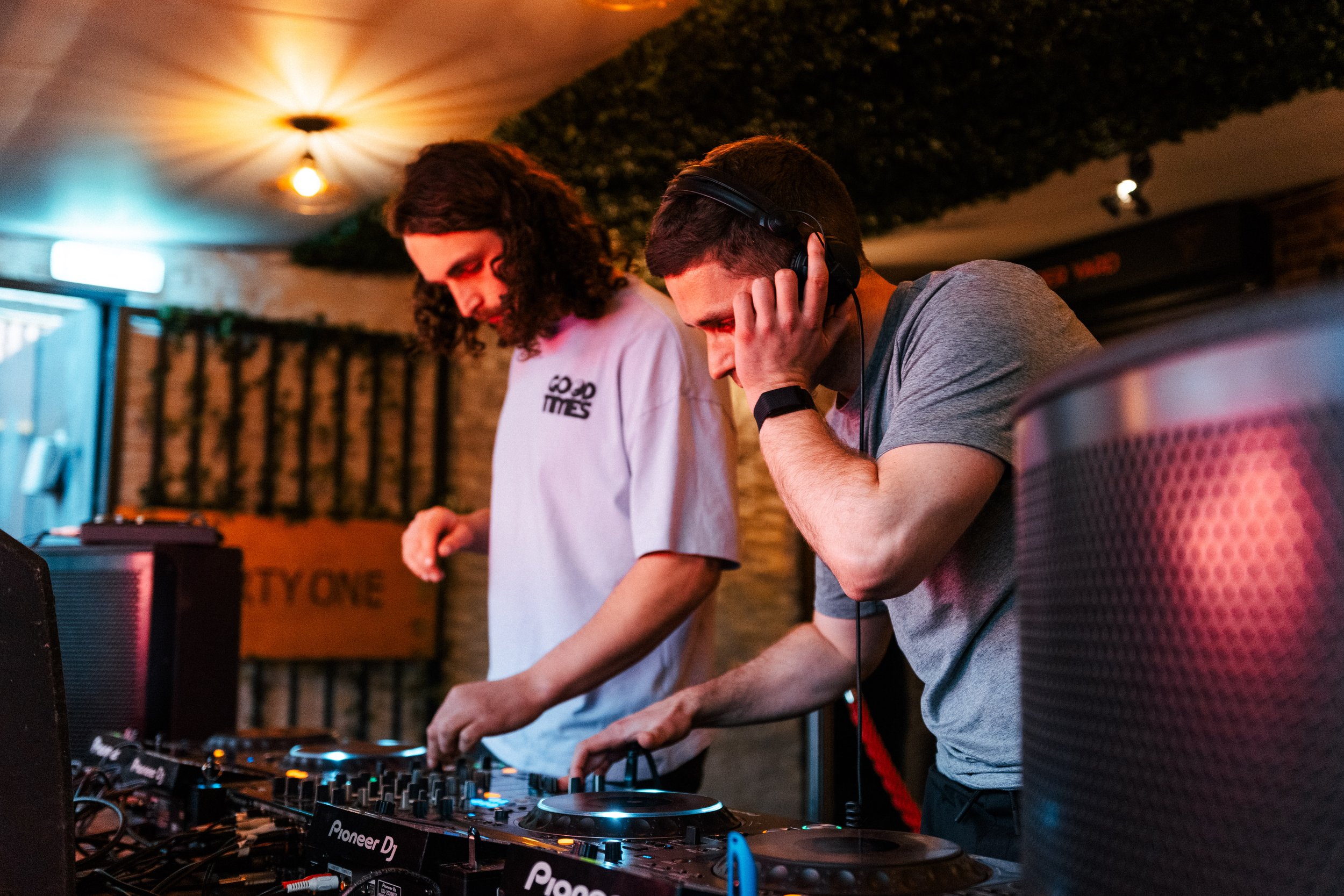 Two young men DJing at a nightclub, one wearing headphones and both focused on the DJ equipment.