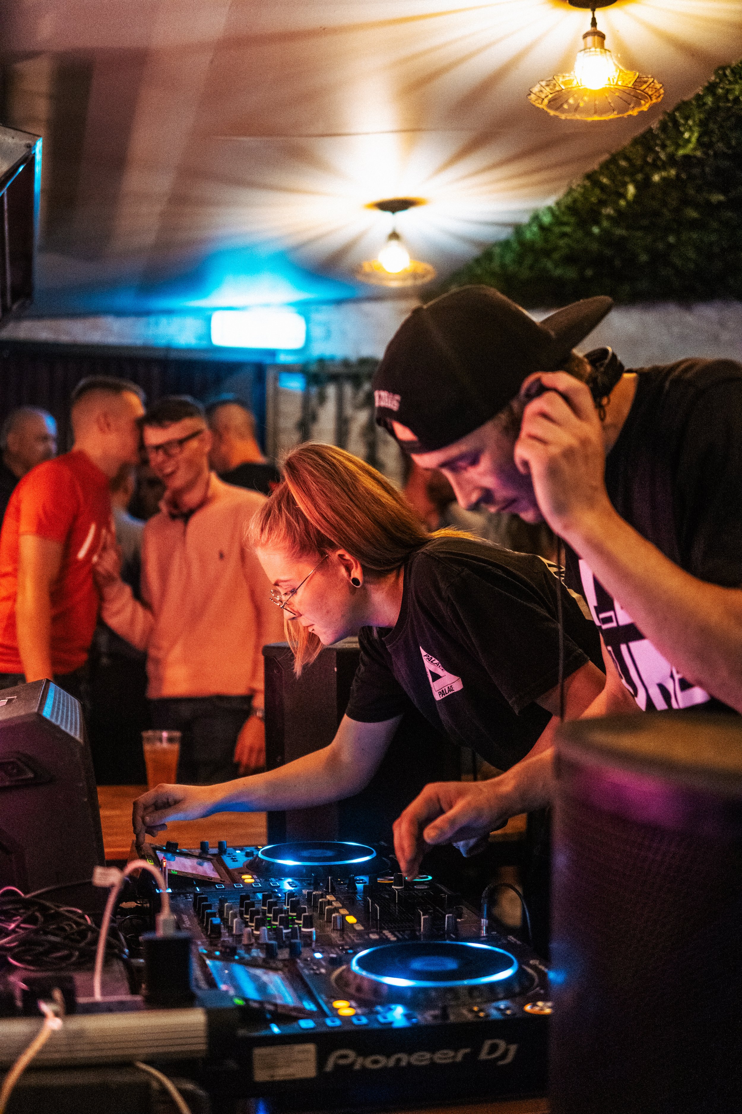 A DJ and a woman are performing at a lively indoor party, with guests mingling and laughing in the background.