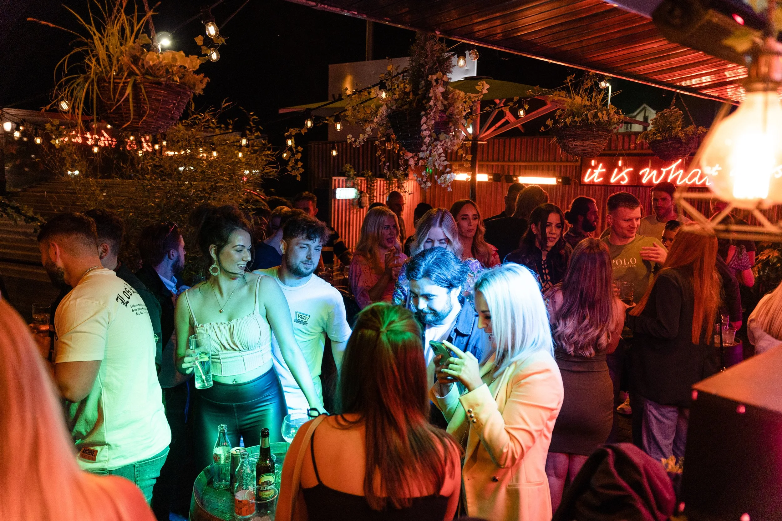 A crowded outdoor party at night with people socializing, drinking, and using their phones under string lights and hanging plants, with a neon sign in the background.
