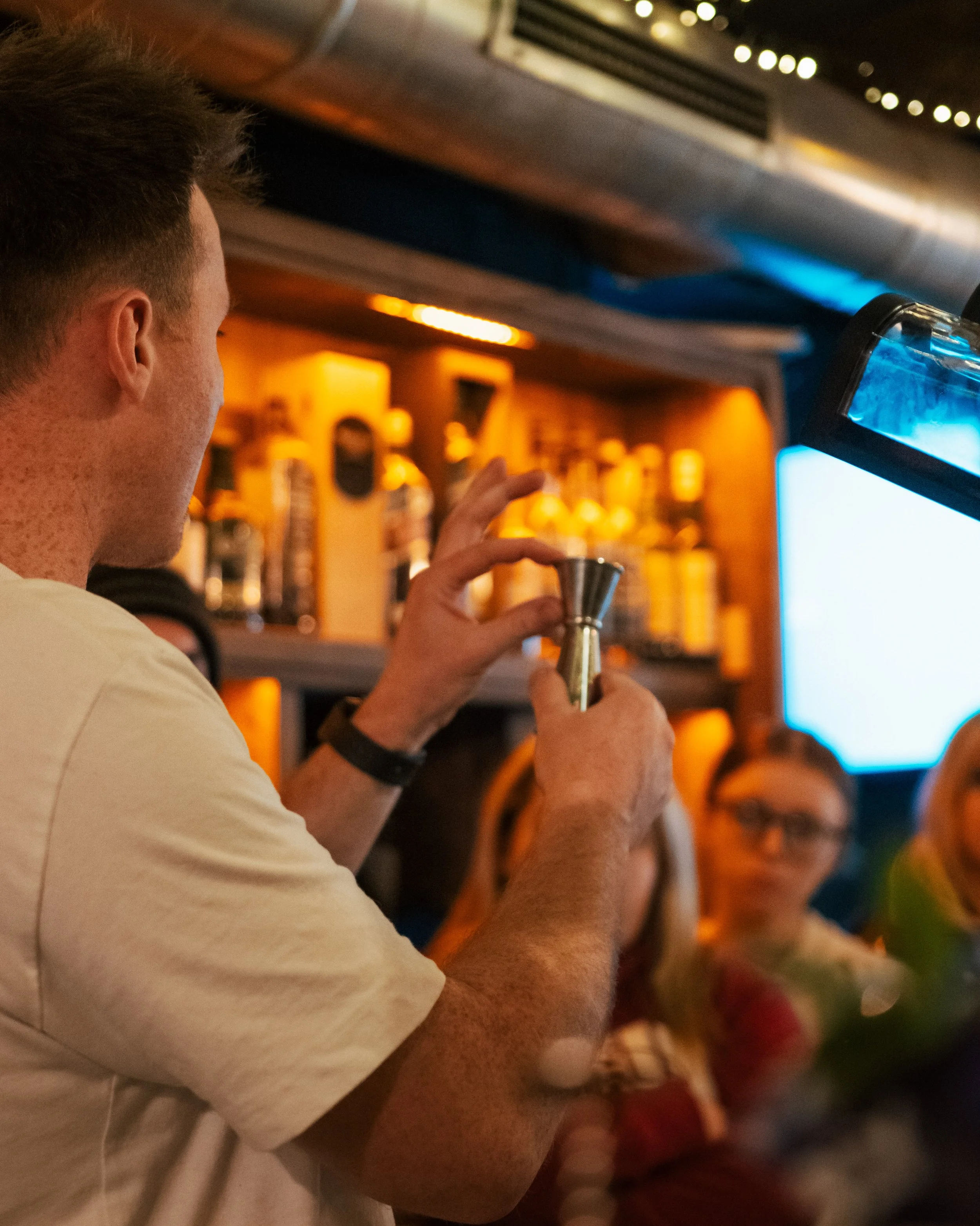 A bartender preparing a drink in a dimly lit bar. The bartender is holding a metal jigger, with shelves of liquor bottles behind him and a large screen to the side.