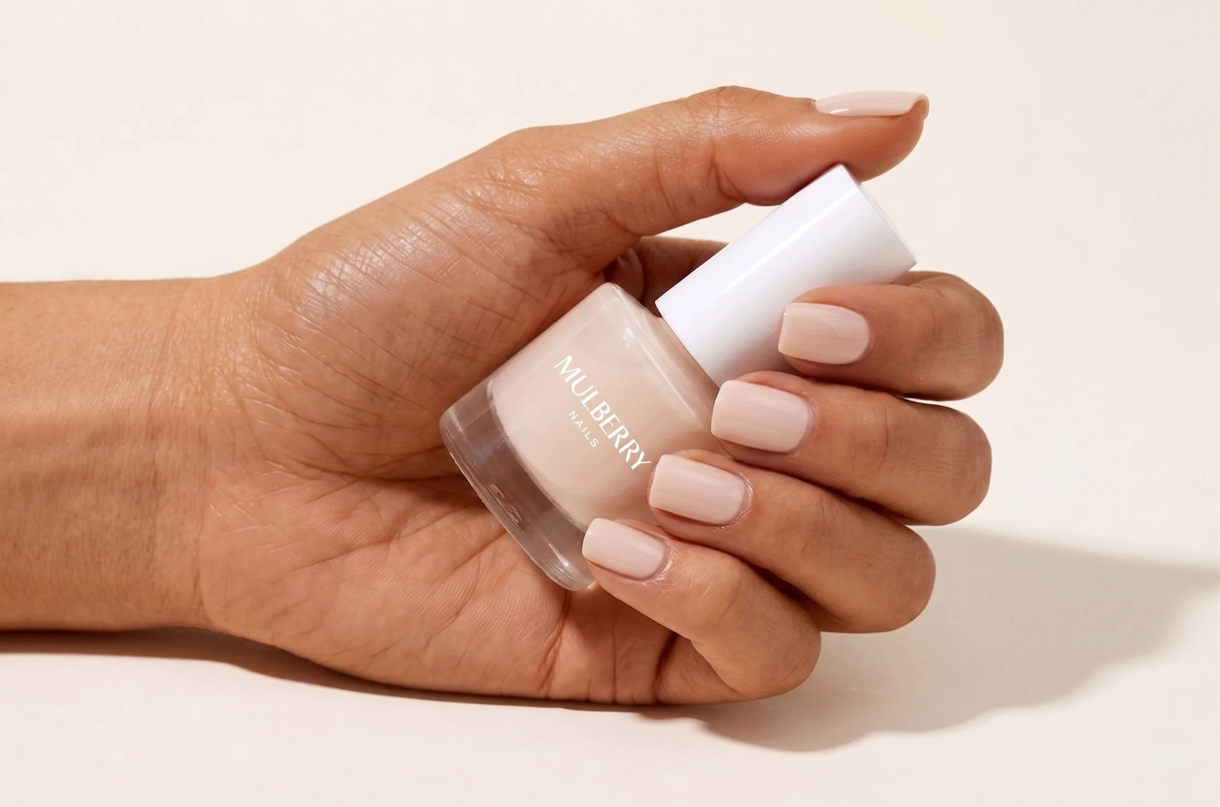 Close-up of a hand holding a bottle of nude-colored nail polish labeled "MULBERRY NAILS," with a plain white background.