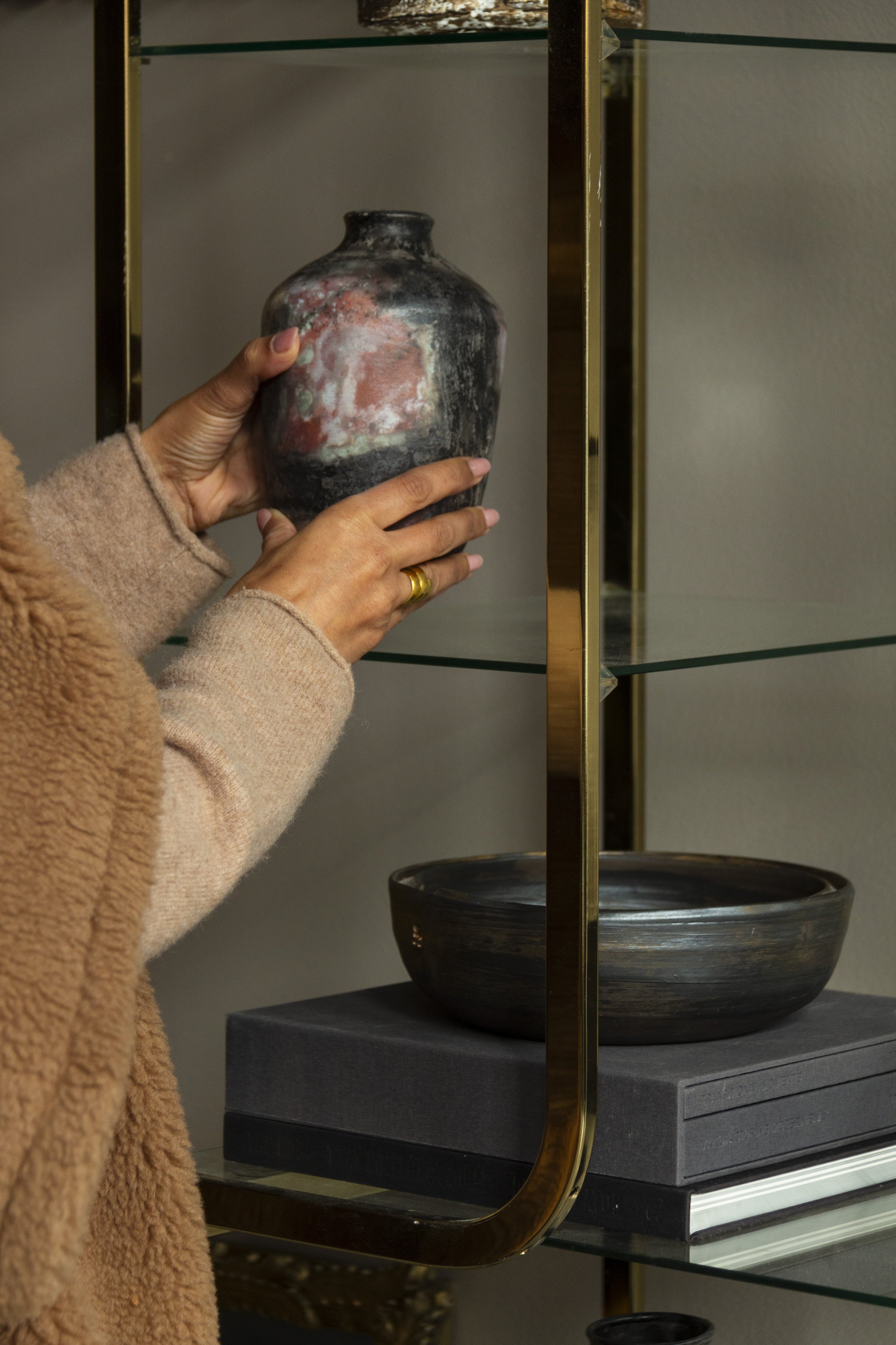 Person placing a rustic vase on a glass shelf with a brass frame, next to a ceramic bowl and stacked books.