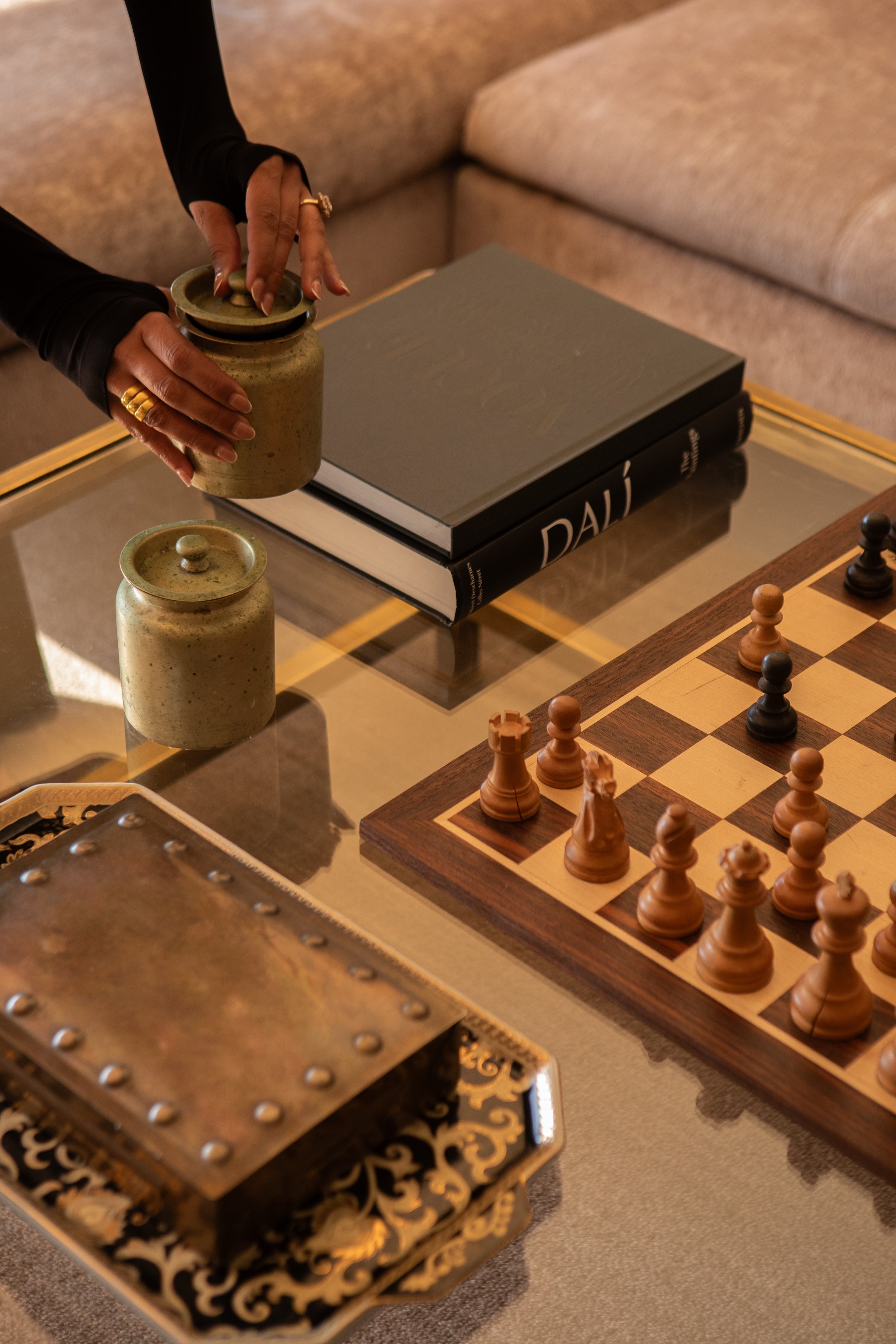 Close-up of a person arranging a decorative jar on a glass table with a chessboard, a book, and a metal decorative box.